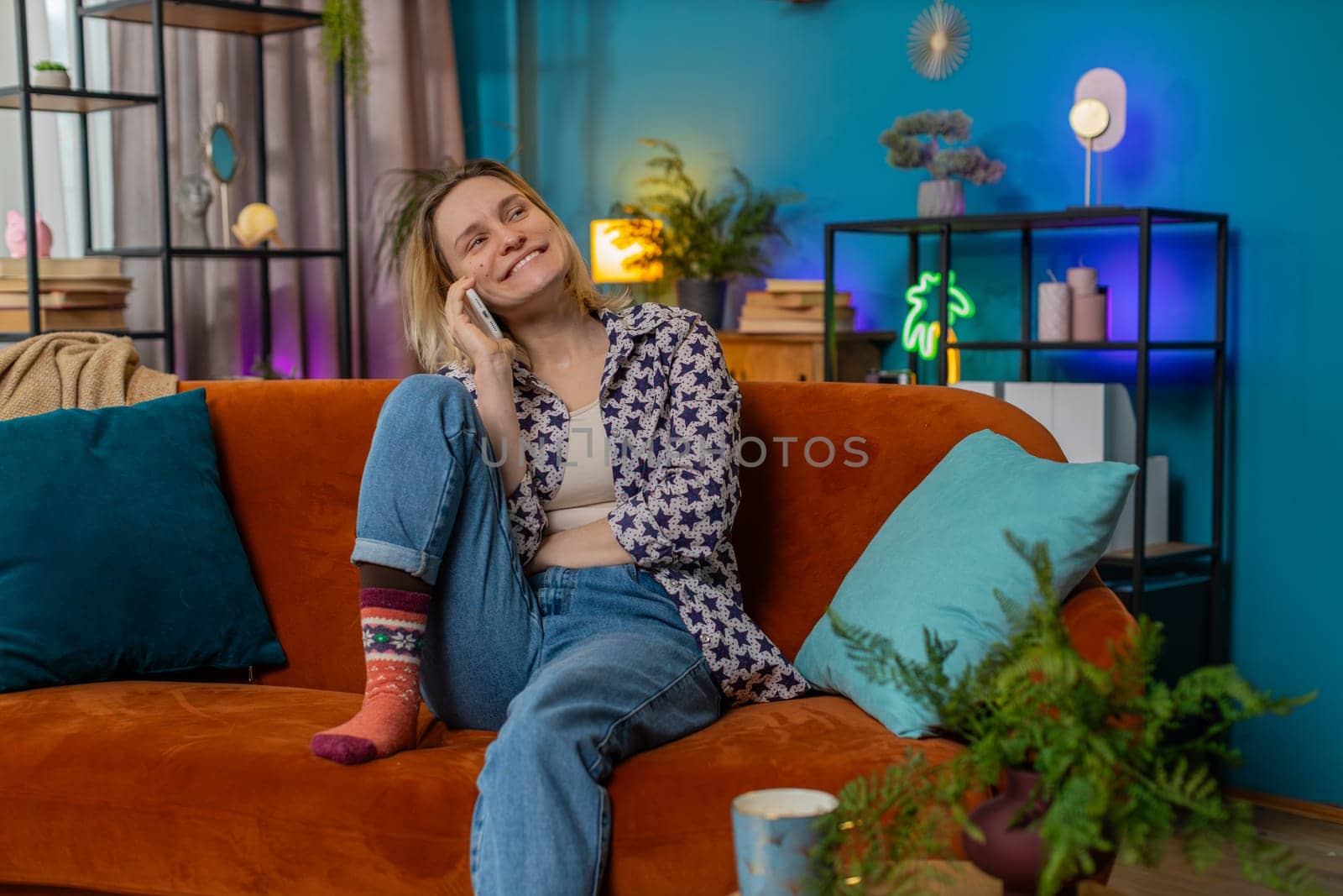 Happy young Caucasian woman making mobile phone conversation with friends sitting on couch at home by efuror