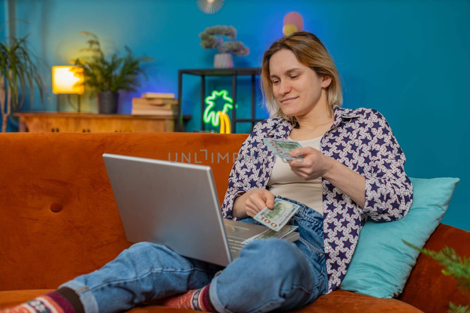 Planning budget. Rich happy young woman freelancer counting money cash use laptop computer calculate domestic bills sitting on sofa at home. Caucasian girl satisfied of income earnings in living room.