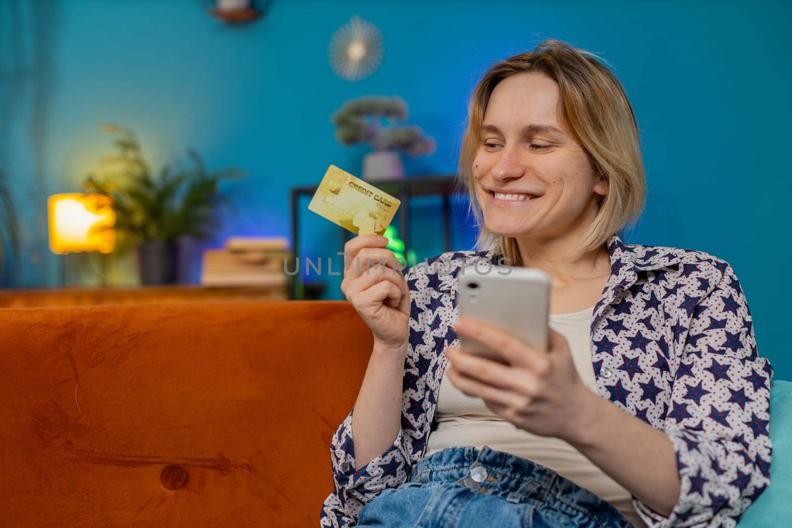 Happy excited woman doing payments online purchase shopping in internet store. Smiling Caucasian girl holding banking credit card involved in verification process in mobile application on sofa at home