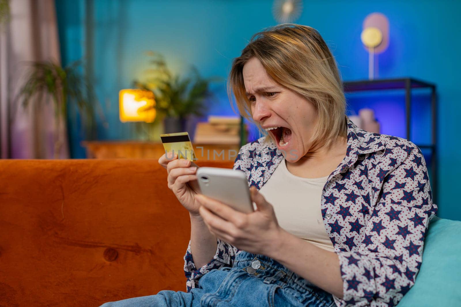 Angry stressed woman trying to pay online shopping with smartphone blocked credit card smartphone. Annoyed Caucasian girl buying bank refuse problem unsuccessful payment lack of money balance at home.