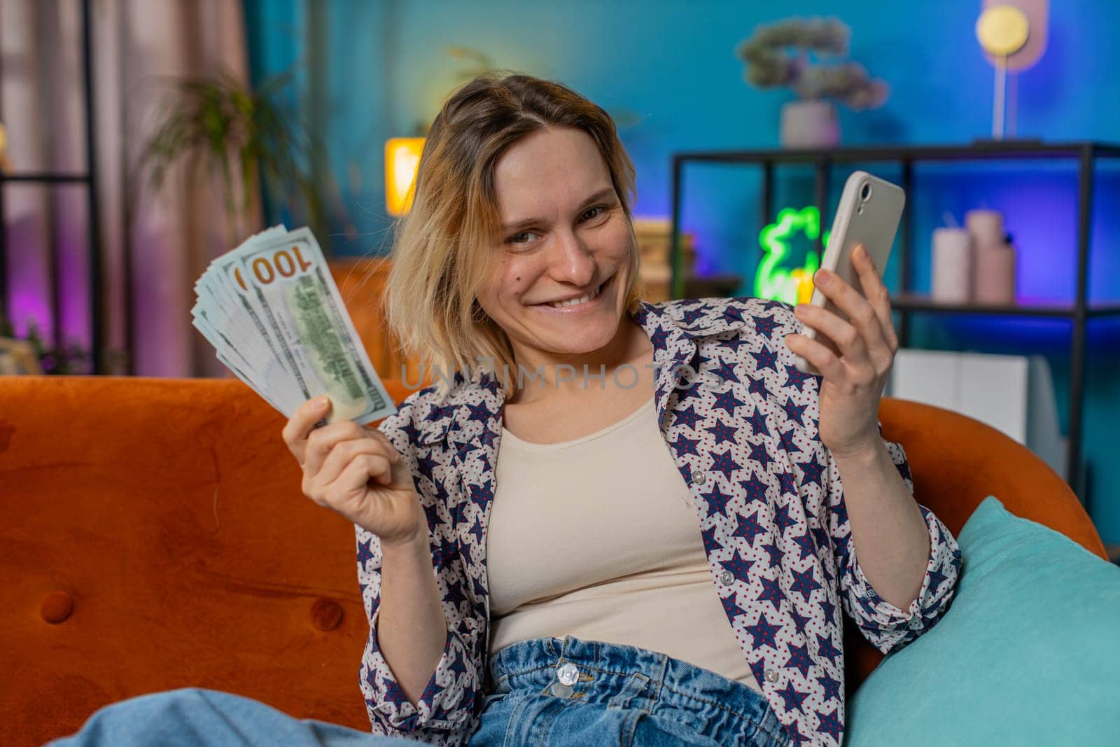 Successful rich smiling woman holding smartphone and waving dollar bill money fan sitting on sofa at home. Happy Caucasian girl winning online lottery game planning vacation calculating budget concept