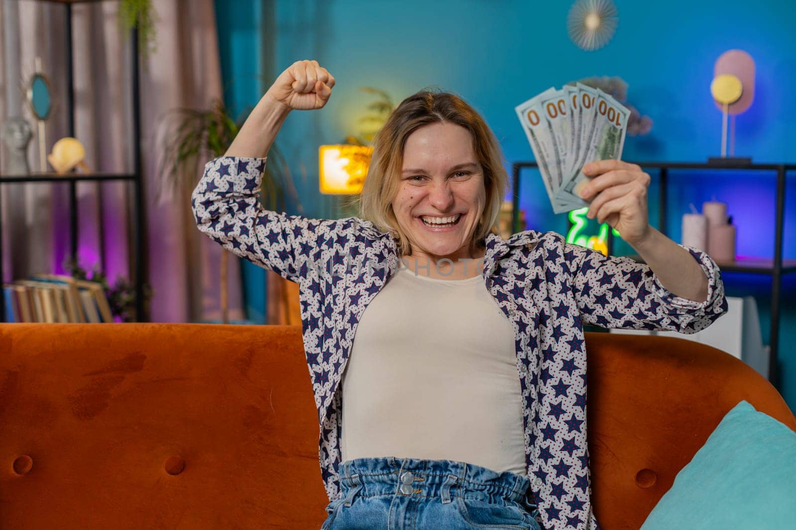 Successful rich blonde woman holding waving dollar bill money fan sitting on couch at home room. Happy excited girl winning online casino lottery game planning vacation calculating budget at home.