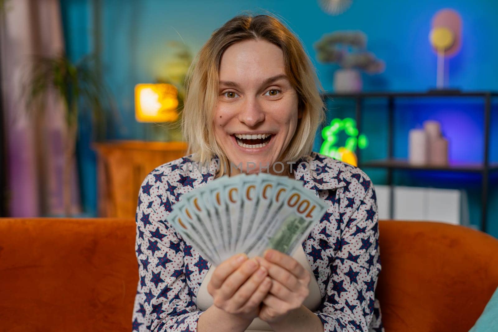 Successful rich blonde woman holding waving dollar bill money fan sitting on couch at home room. Happy excited girl winning online casino lottery game planning vacation calculating budget at home.