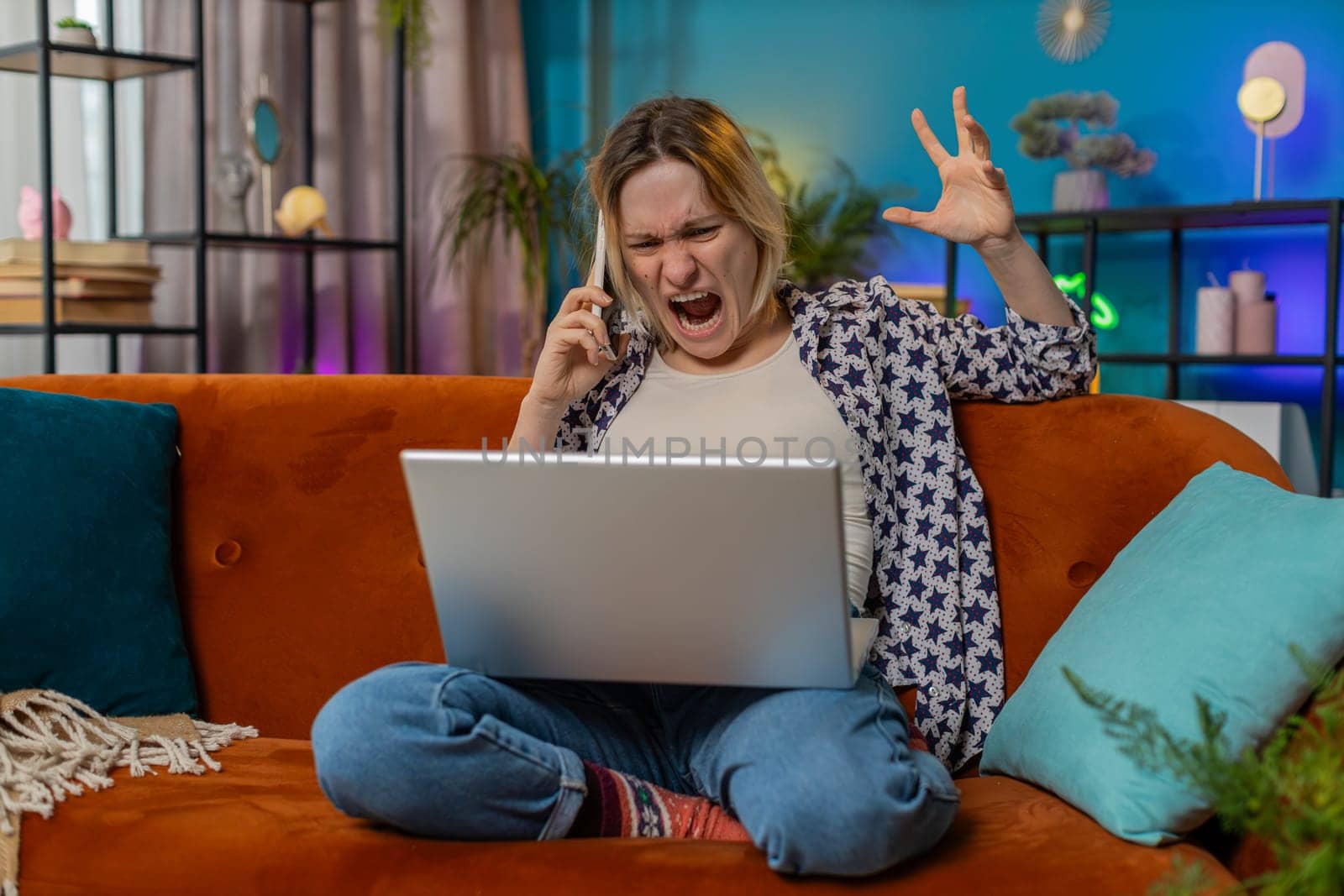 Quarrel. Furious annoyed young woman freelancer engage in heated phone conversations sitting on sofa. Upset Caucasian blonde girl expresses frustration using cellphone and laptop on couch in apartment