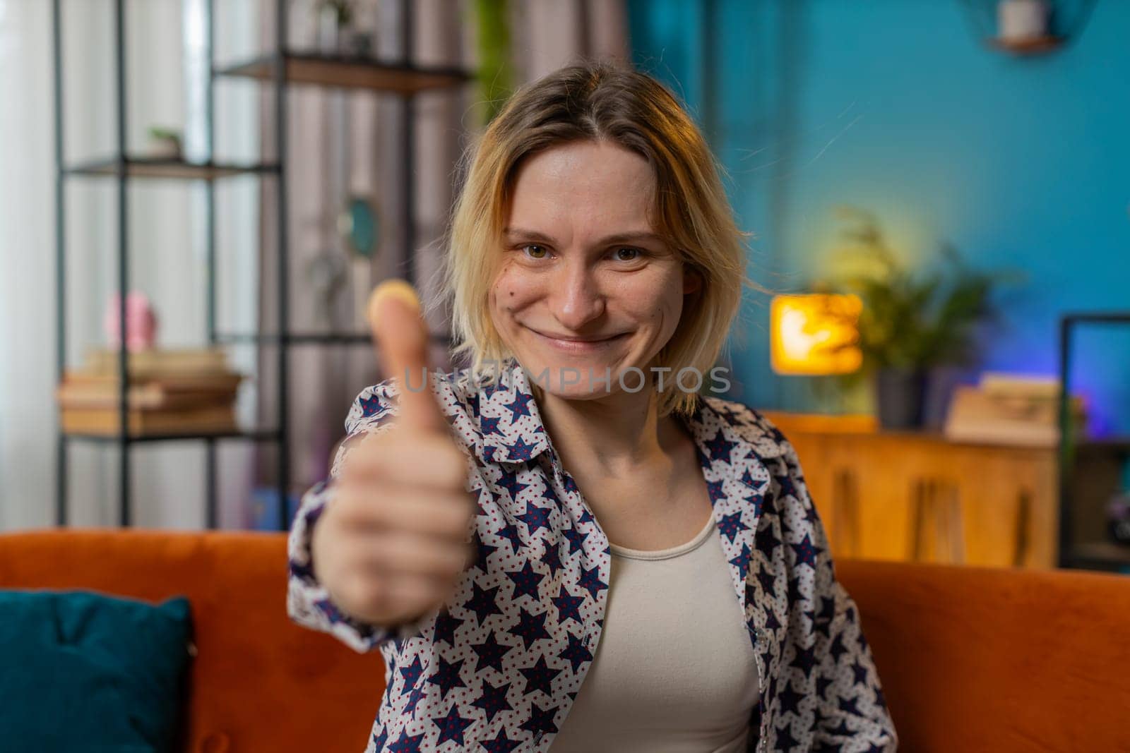 Portrait of excited smiling Caucasian woman looking approvingly at camera showing double thumbs up, like sign, good news, positive feedback. Happy young girl sitting on sofa in living room at home.