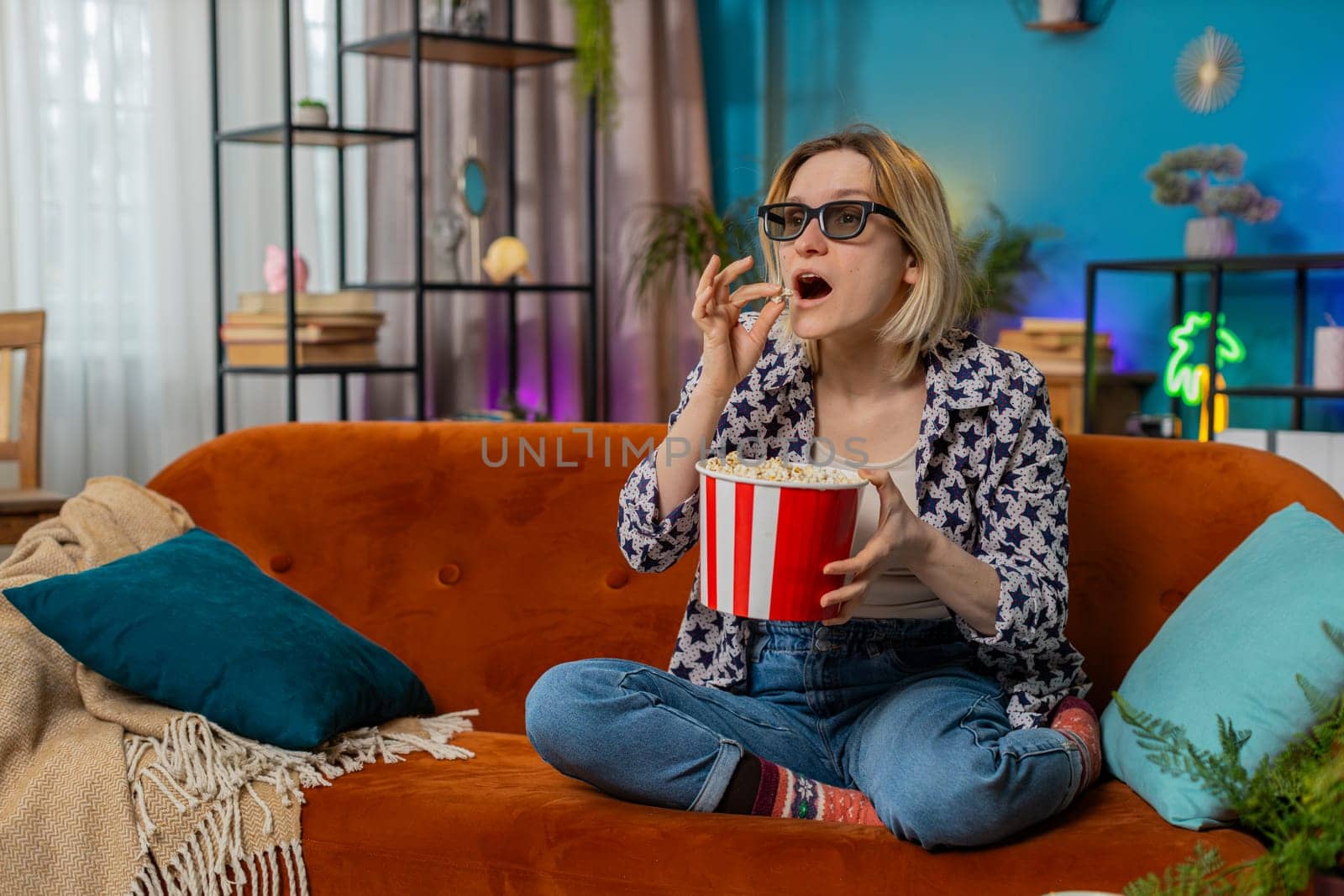 Excited young woman sitting on couch eating popcorn and watching interesting TV serial, sport game, film, online social media movie content at home. Girl in 3D glasses enjoying domestic entertainment.