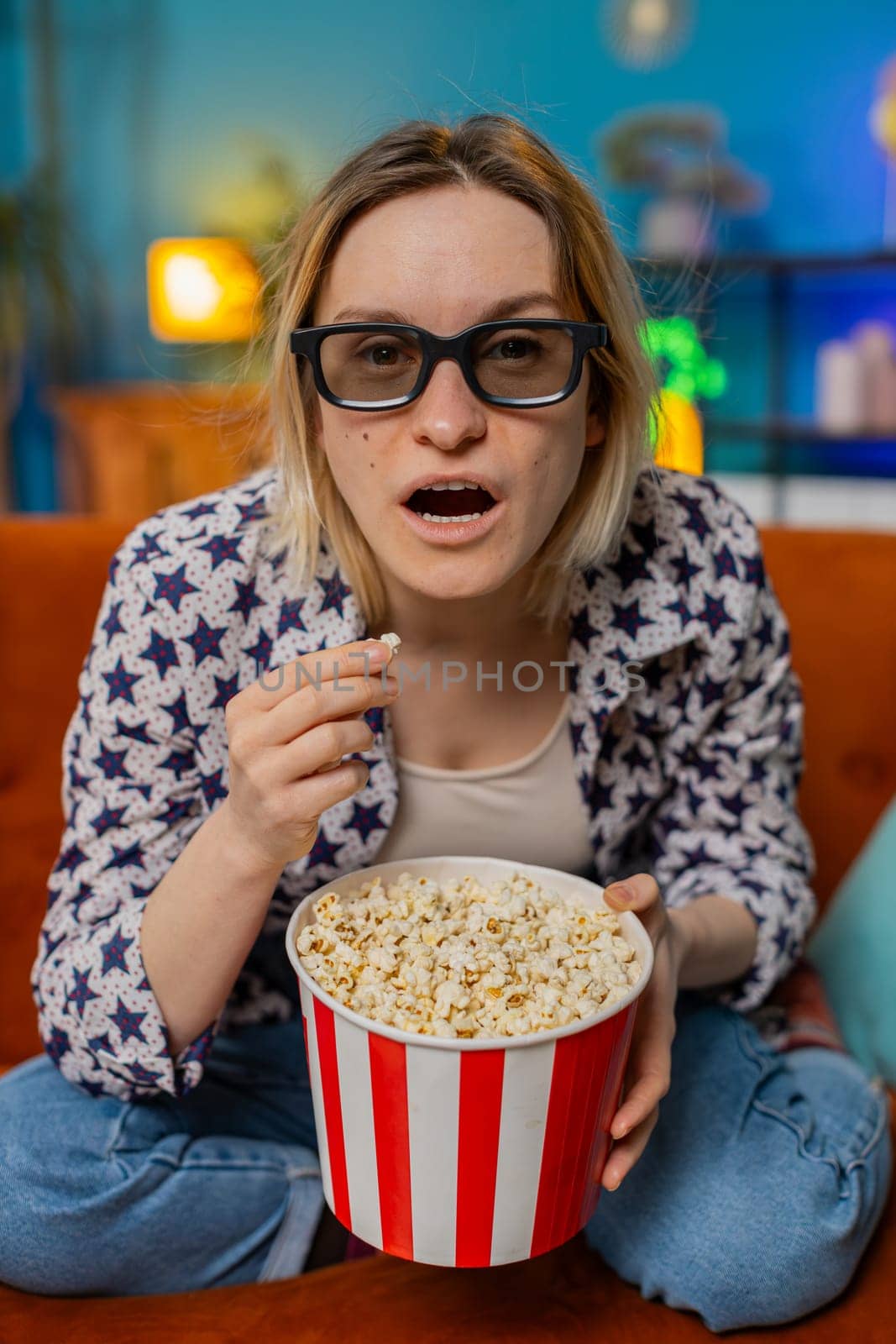 Excited woman sitting on couch eating popcorn watching interesting TV serial, sport game, film, online social media movie content at home. Girl in 3D glasses enjoying domestic entertainment. Vertical