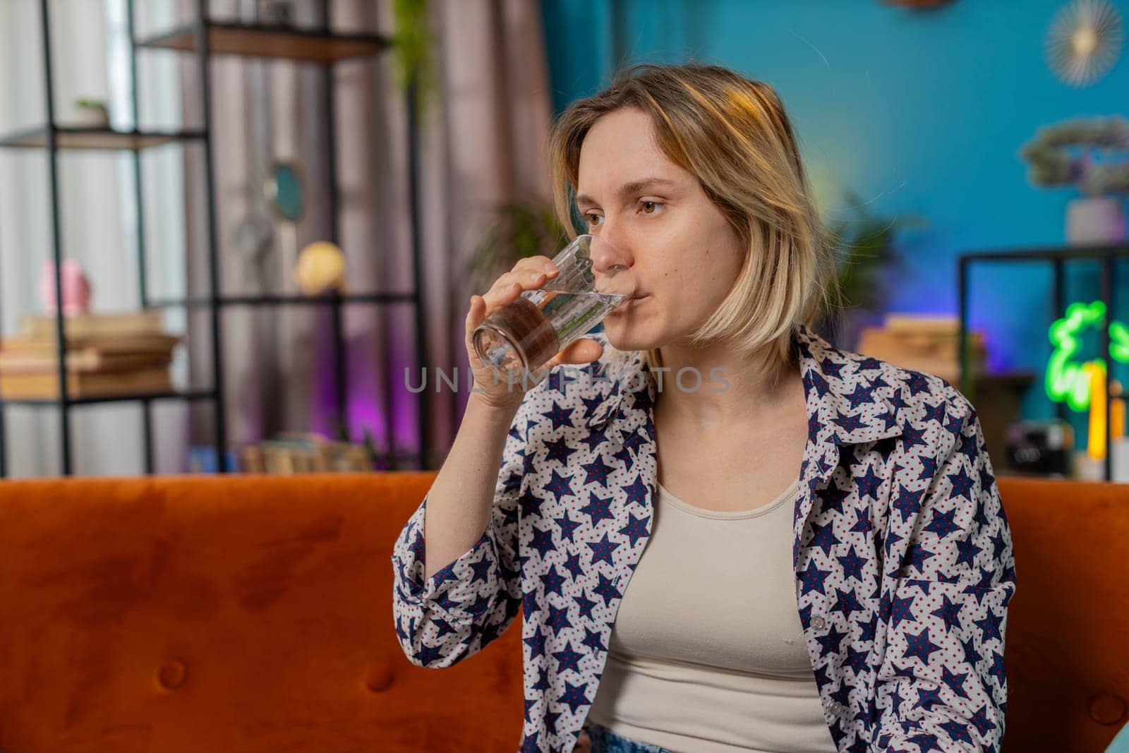 Satisfied young Caucasian woman sitting at home on sofa couch leading healthy lifestyle, drinking a glass of pure water, smiling. Drink daily norm of water. Girl smiling at camera in apartment home.