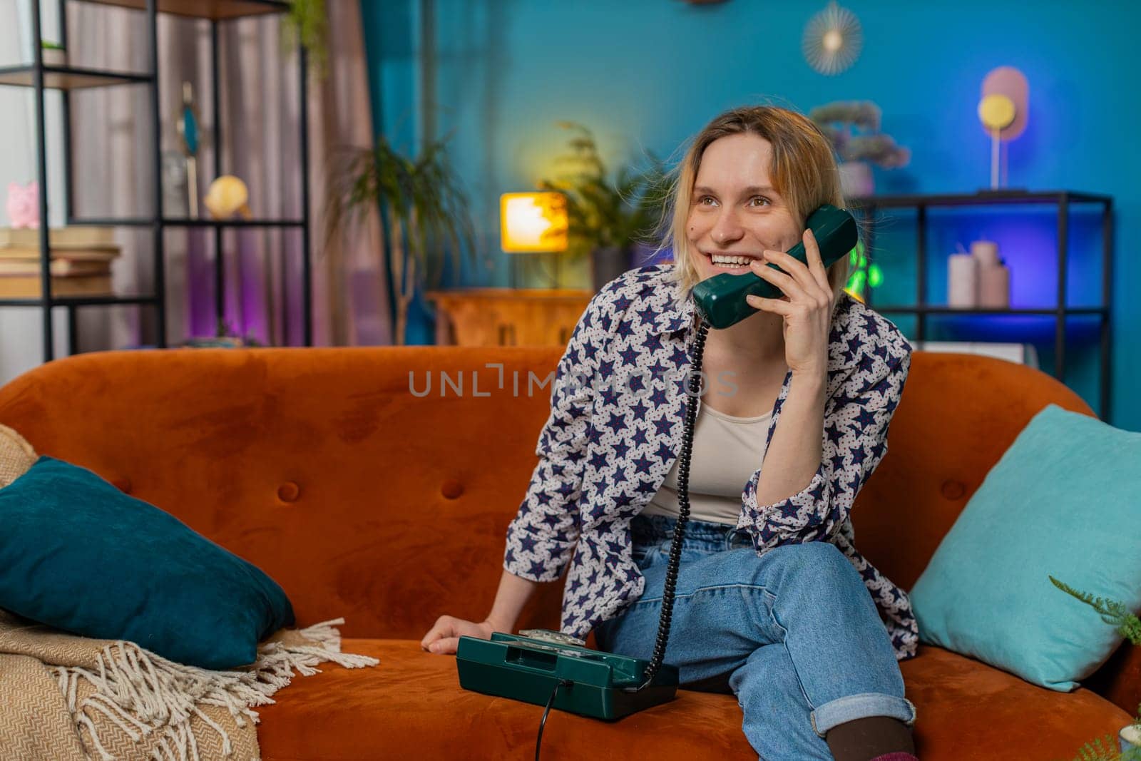 Smiling young woman making wired telephone call conversation with friends sitting on home couch by efuror