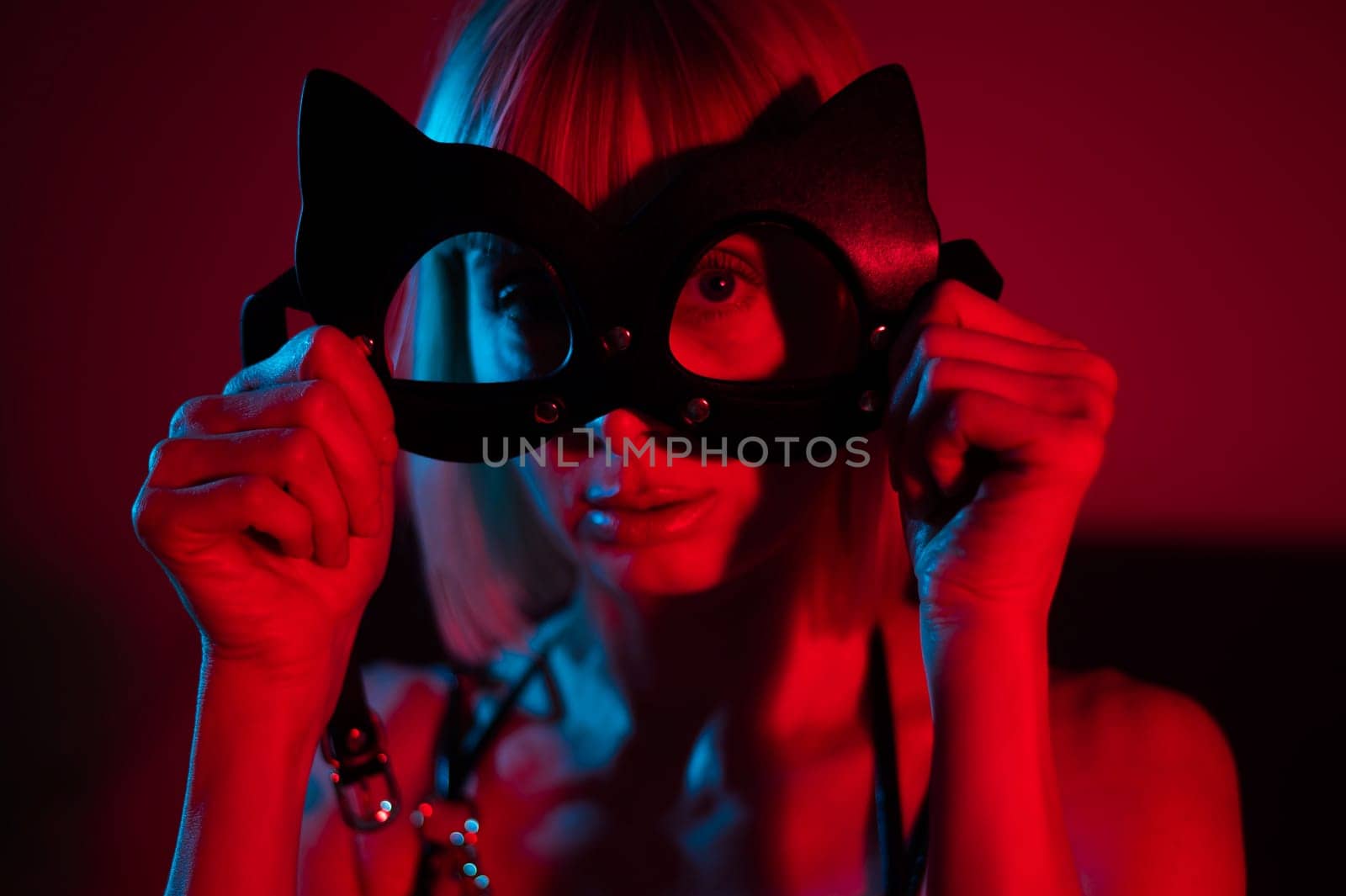 Blonde woman putting on leather mask in bedroom in red blue neon light. by mrwed54
