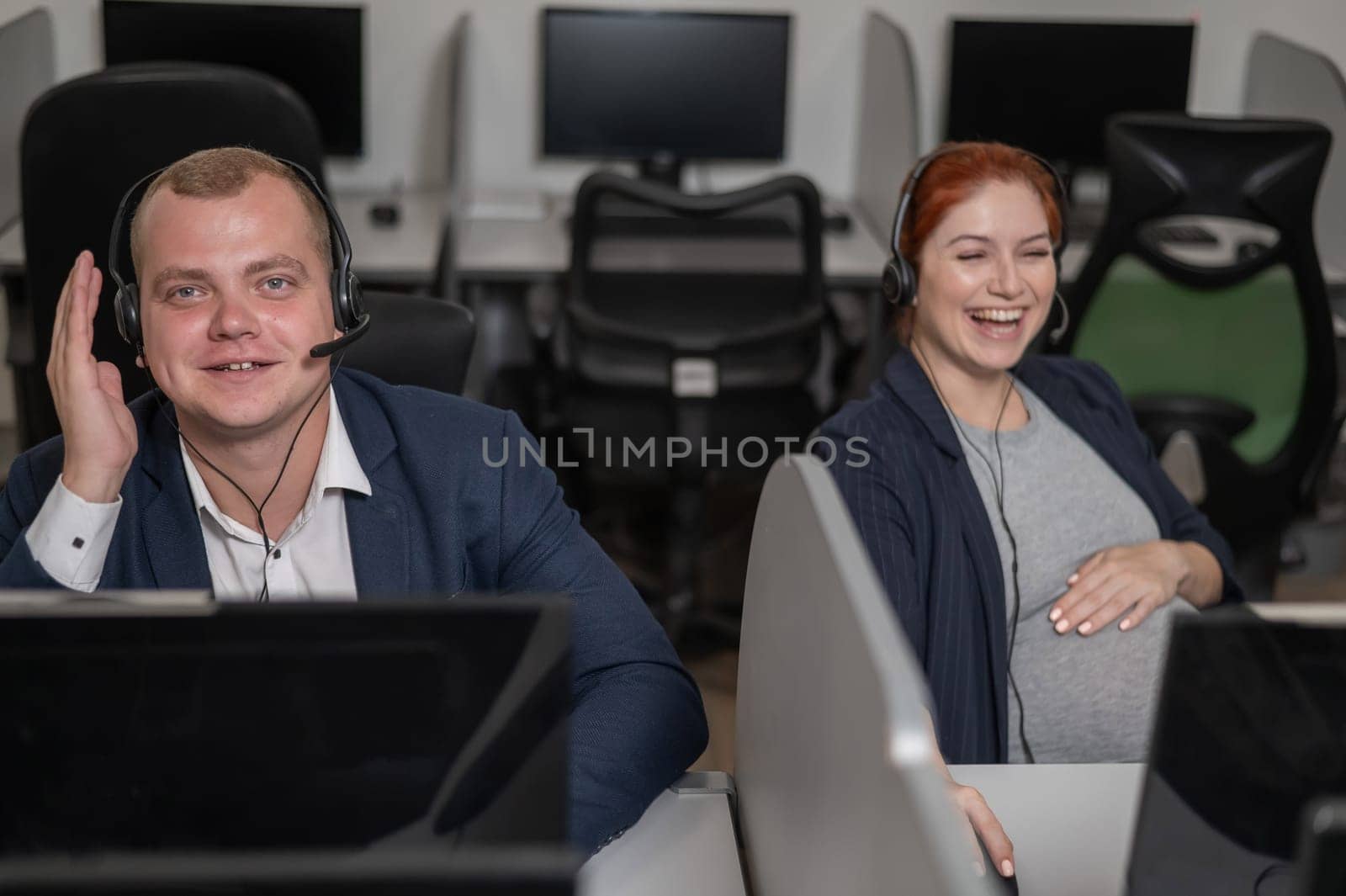 Two friendly call center employees serve customers. Man and pregnant woman talking on a headset in the office