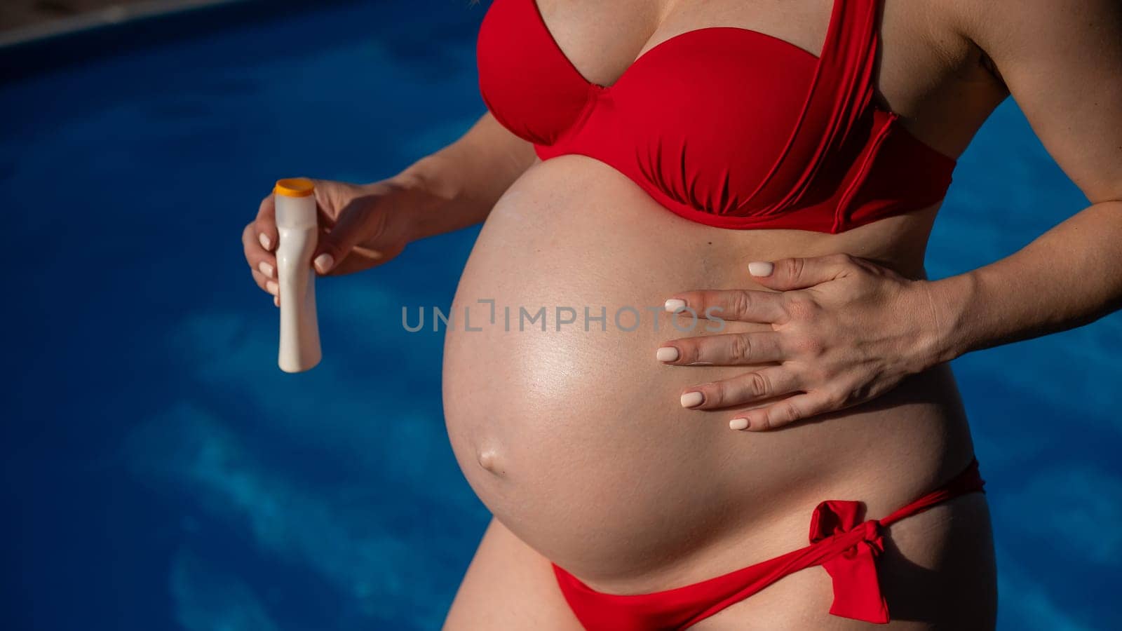 Close-up of the belly of a pregnant woman sunbathing in a red bikini. Expectant mother applies sunscreen