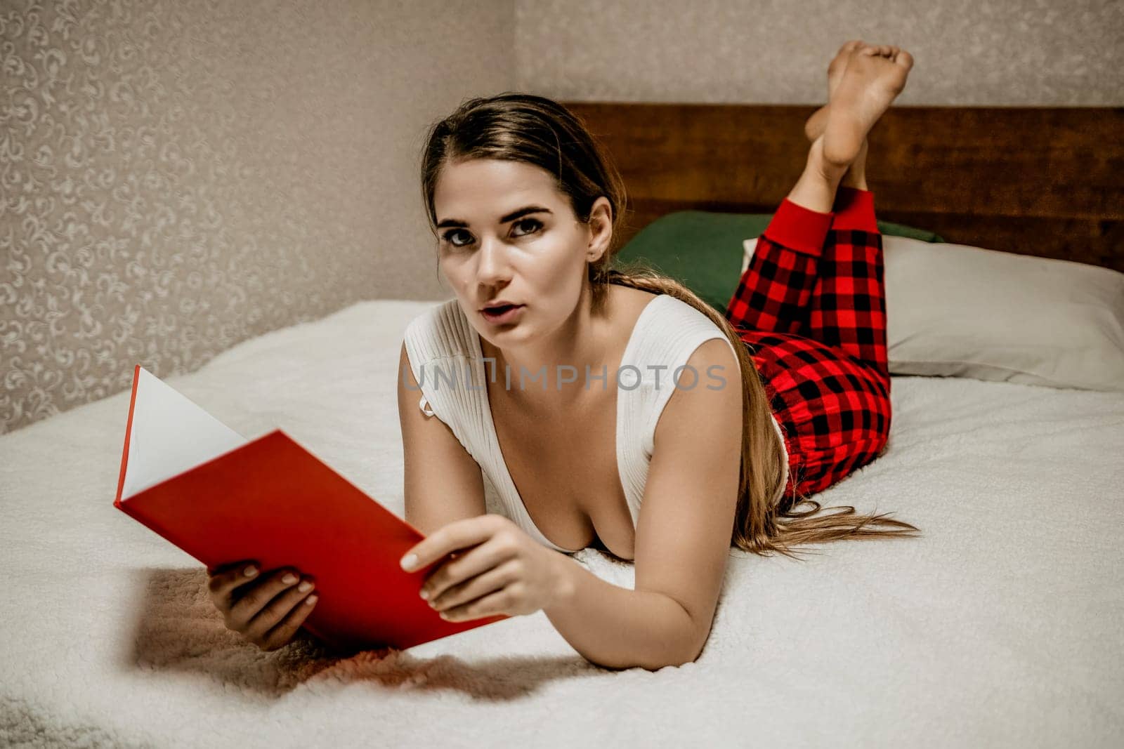 Young brunette woman works and studies from home on a laptop, while lying on her bed in red checkered pants. Balancing work and studies at home