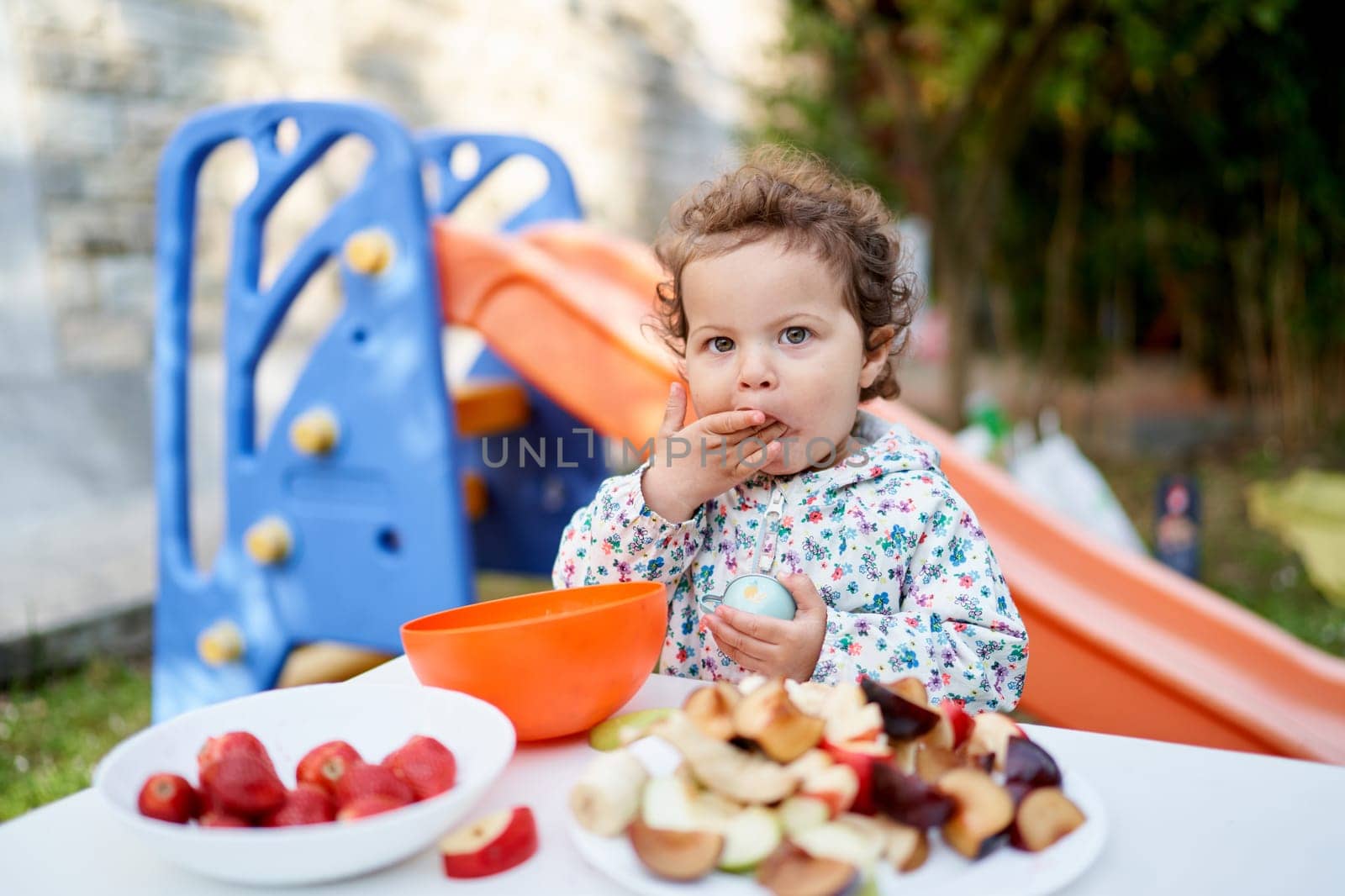 Little girl eats while standing in front of a set table with cut fruits on plates in the garden. High quality photo