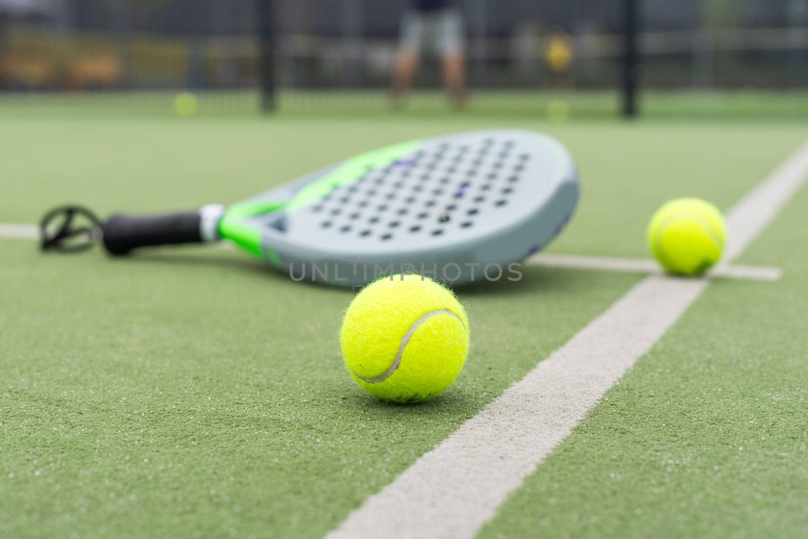 Yellow balls on grass turf near padel tennis racket behind net in green court outdoors with natural lighting. Paddle is a racquet game. Professional sport concept with copy space. by Andelov13