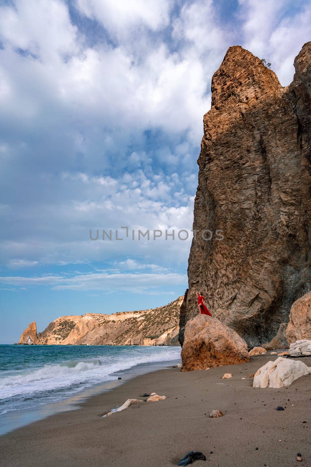 woman sea red dress. Woman with long hair on a sunny seashore in a red flowing dress, back view, silk fabric waving in the wind. Against the backdrop of the blue sky and mountains on the seashore