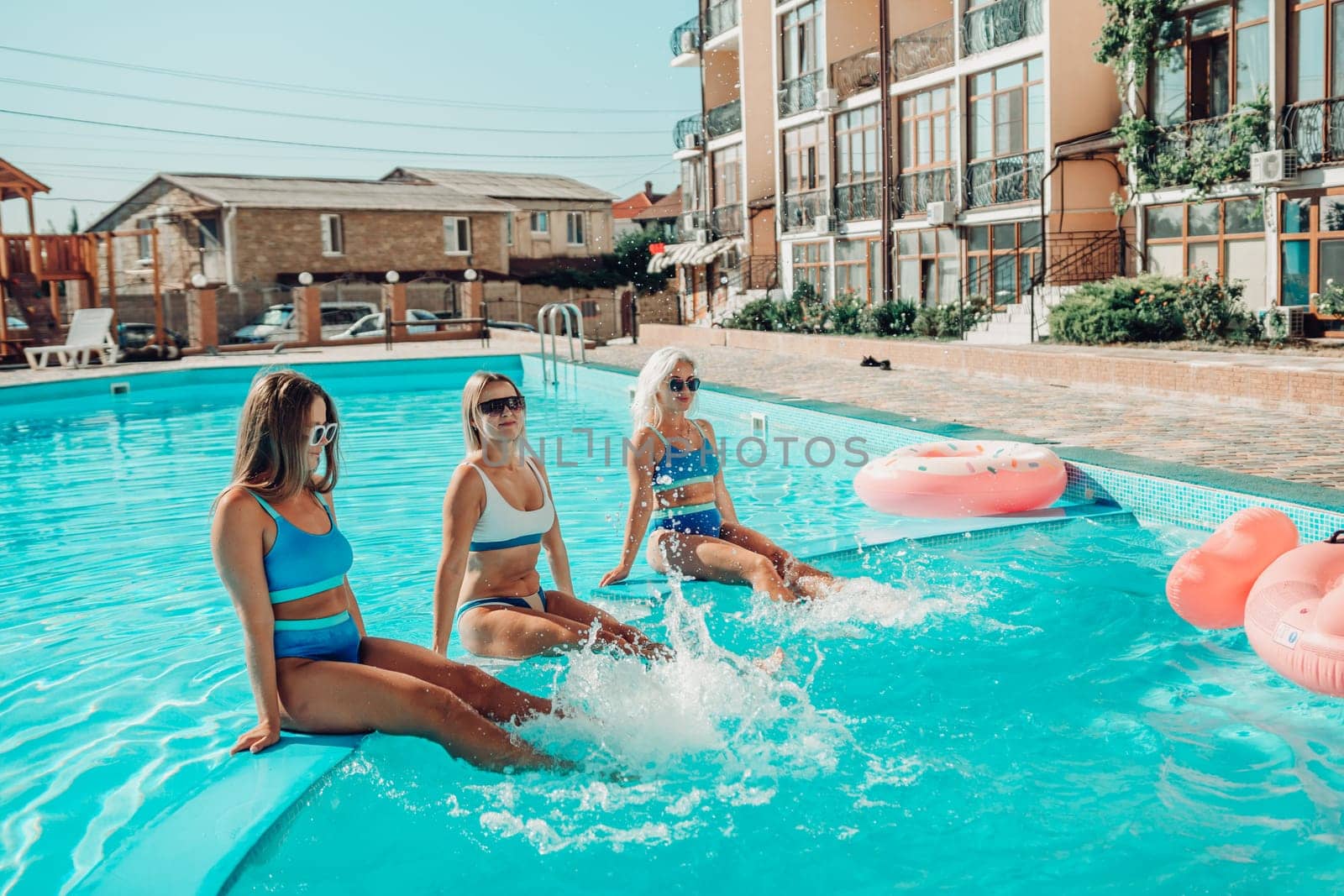 Three women are sitting in a pool with pink and blue floaties. Scene is lighthearted and fun