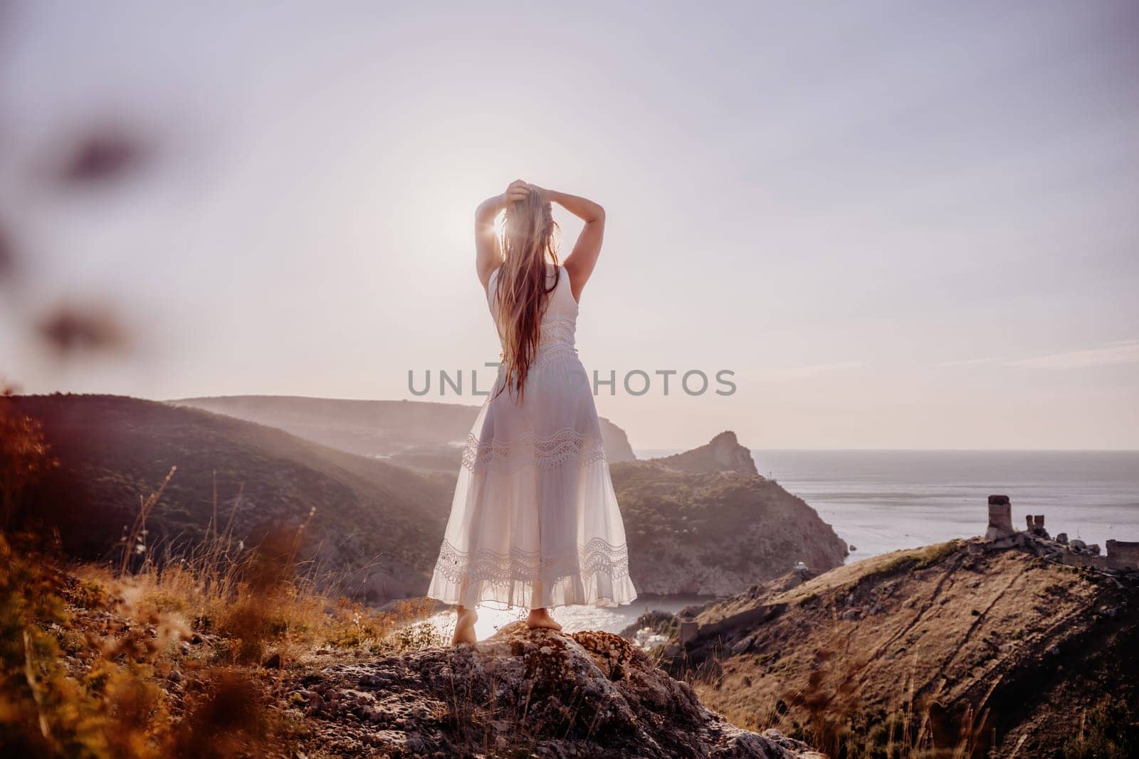 A woman stands on a hill overlooking the ocean. She is wearing a white dress and has her hands on her head. The scene is serene and peaceful, with the woman looking out over the water. by Matiunina