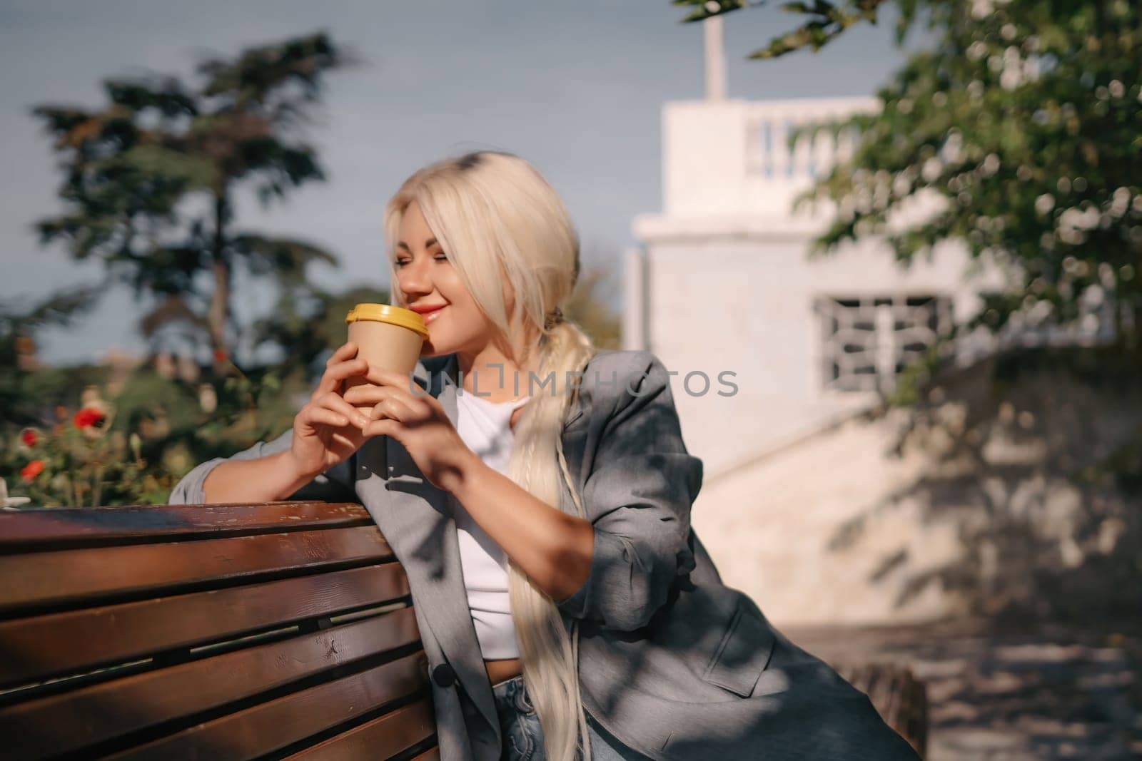 A blonde woman is sitting on a bench with a cup of coffee in her hand. She is smiling and looking out into the distance