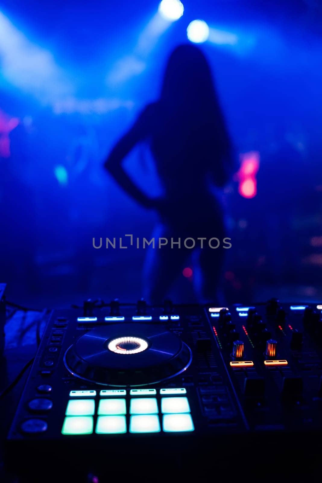 silhouette of young slim dancers on the stage out of focus through the DJ booth and a mixer with a blue background