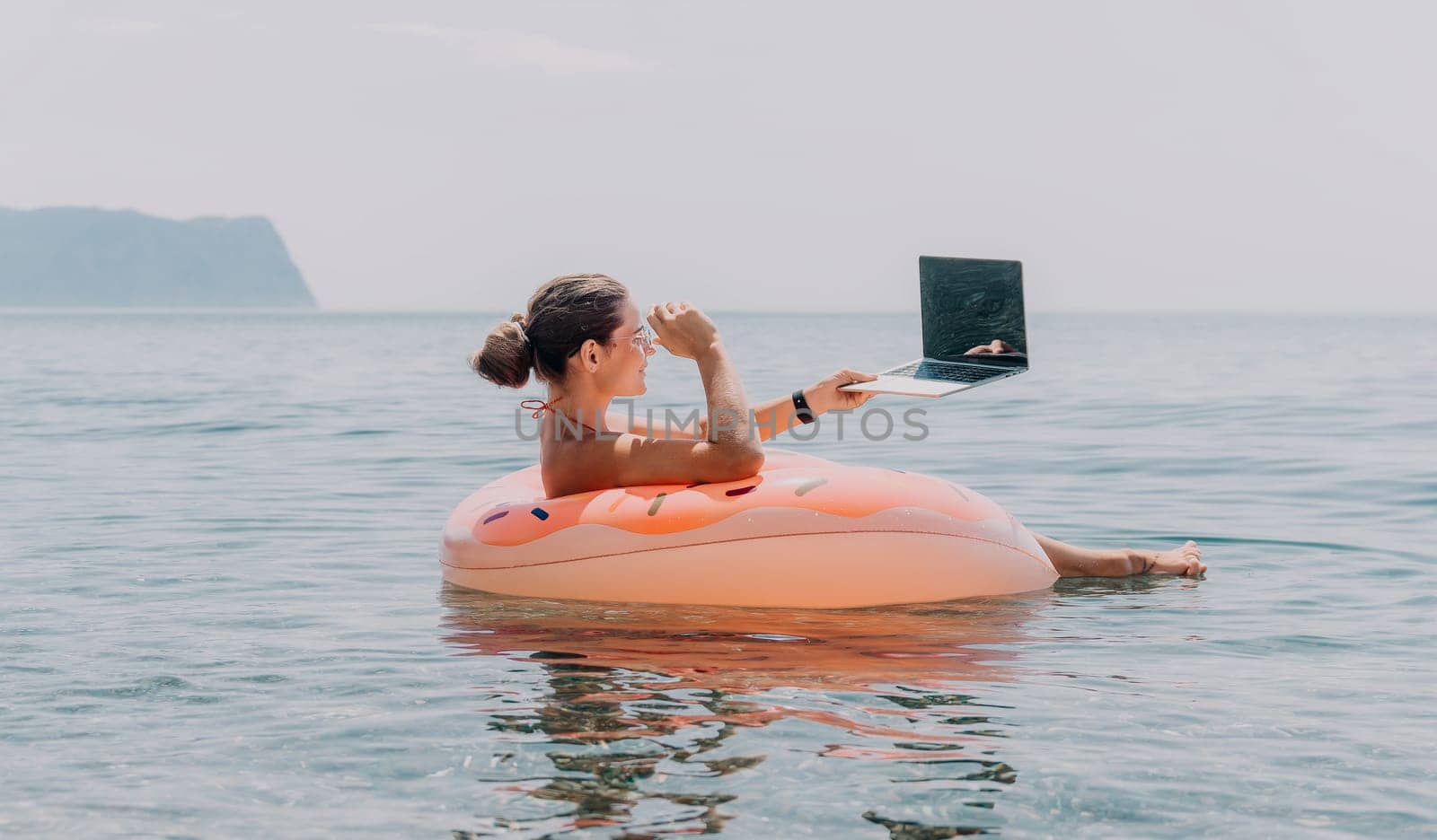 Woman freelancer works on laptop swimming in sea on pink inflatable ring. Happy tourist floating on inflatable donut and working on laptop computer in calm ocean. Freelance, remote working anywhere by panophotograph