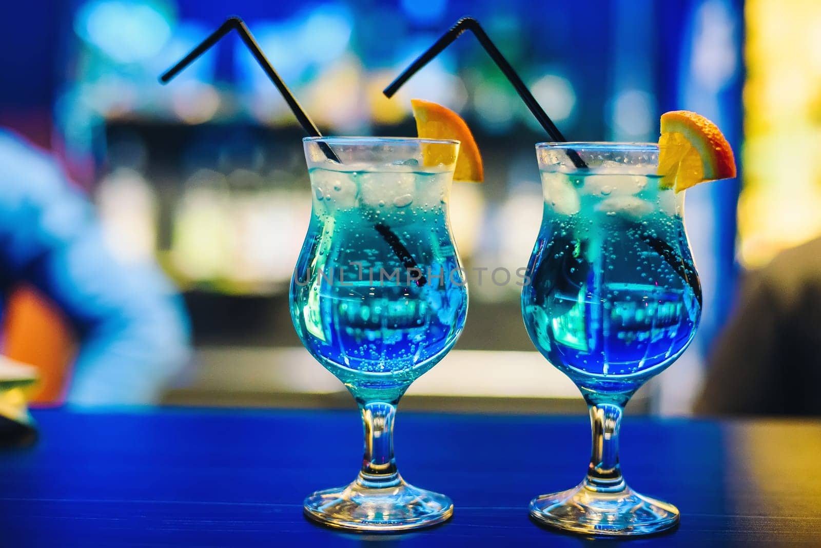 two glasses of blue cocktail Martini with orange and ice cubes and straws on the bar