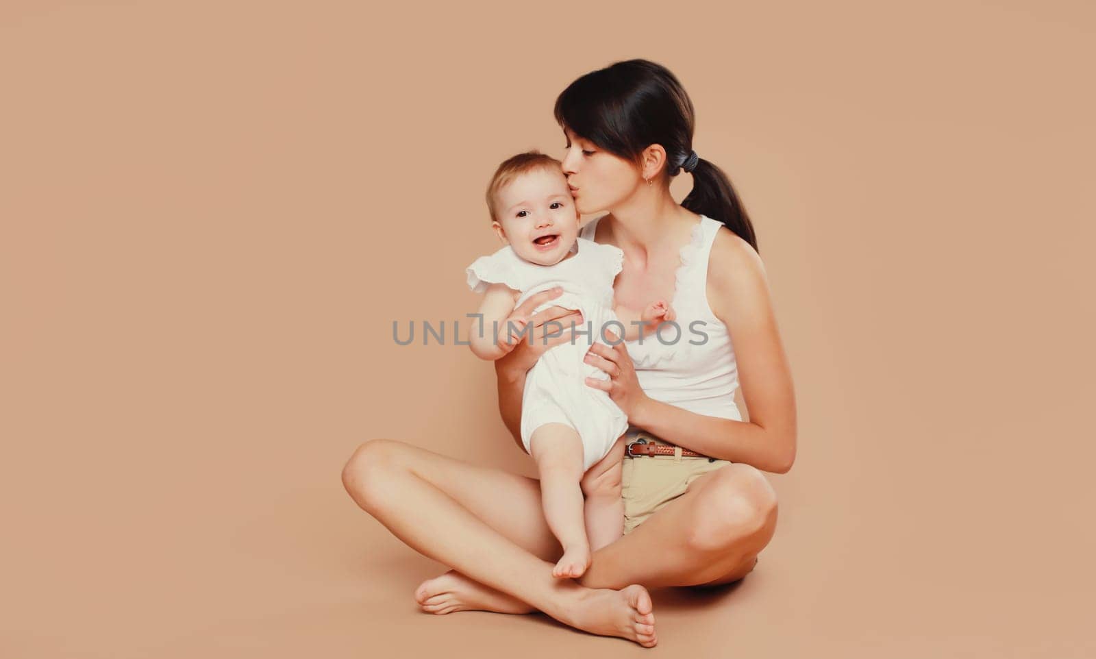 Happy young mother holding and kissing baby on studio background by Rohappy