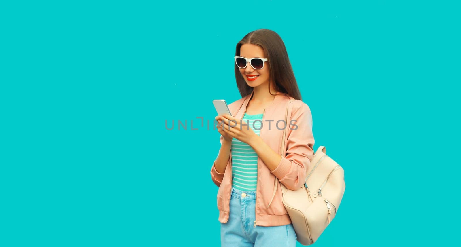 Portrait of smiling young woman with smartphone wearing backpack and sunglasses on blue background