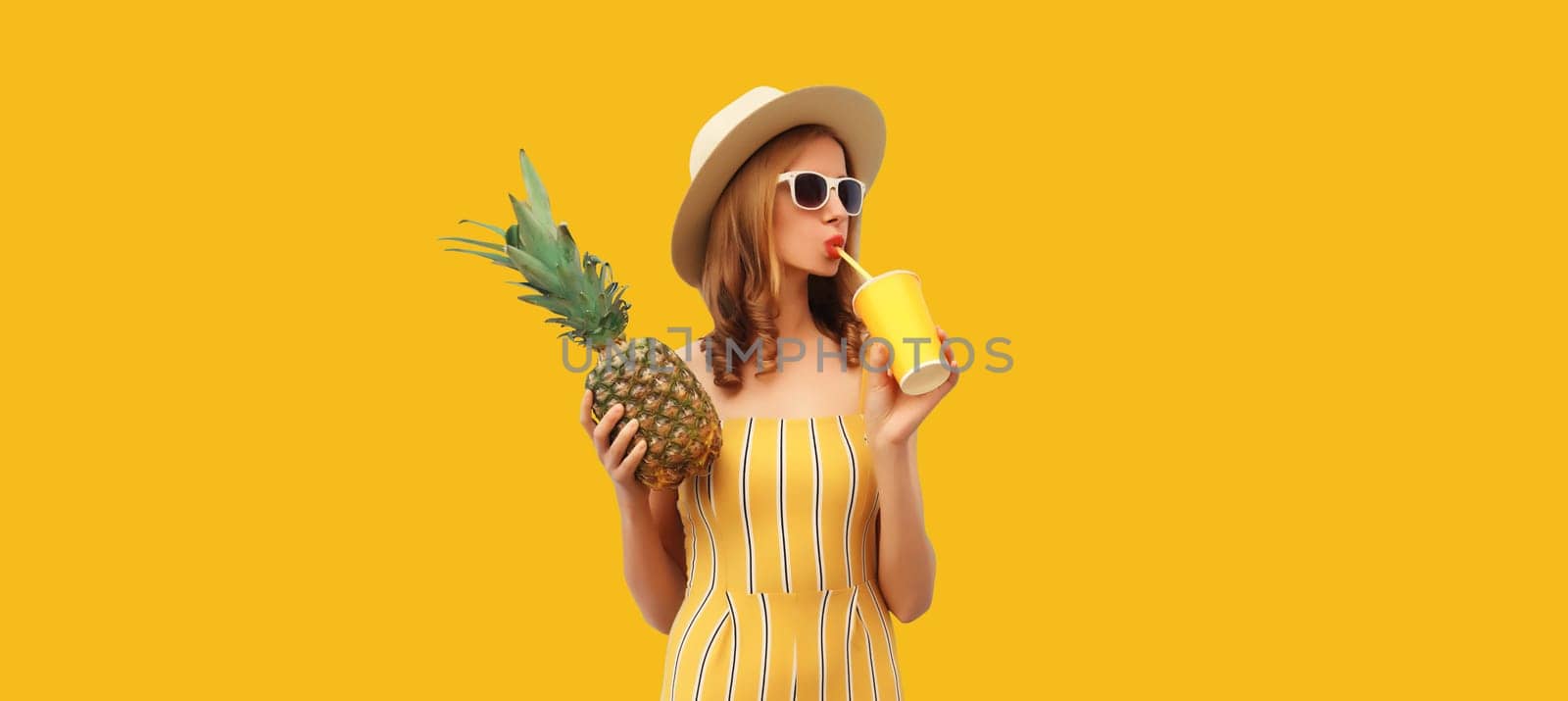 Summer bright portrait of stylish young woman drinking fresh juice holding pineapple fruit in straw hat, sunglasses on colorful yellow studio background