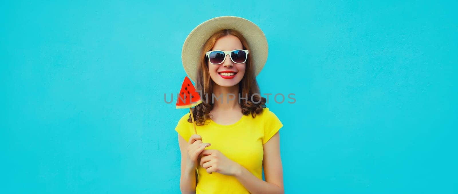 Summer portrait of happy young woman with lollipop or ice cream shaped slice of watermelon wearing straw hat on blue studio background