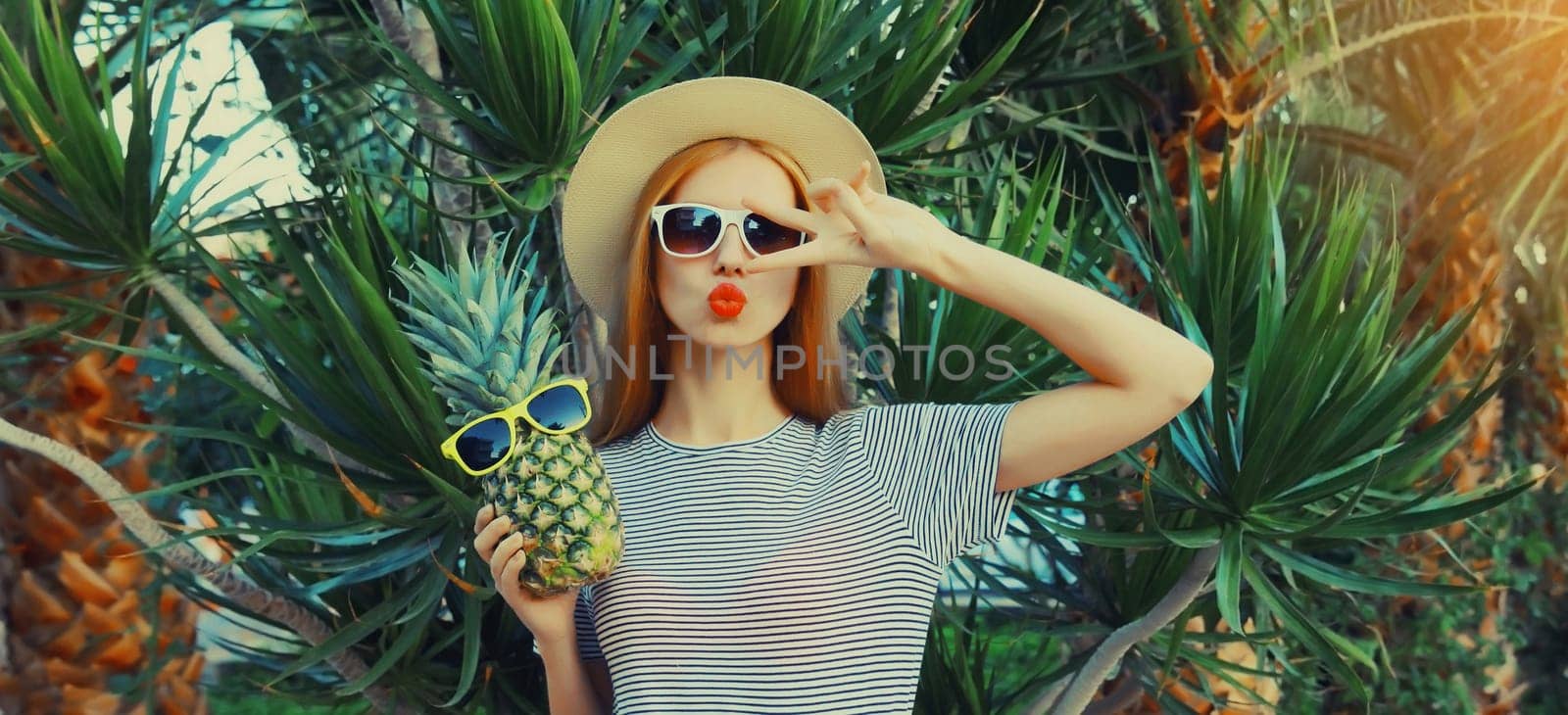 Summer portrait of beautiful young woman with pineapple fruits posing blowing lips sends kiss wearing sunglasses, straw hat on palm tree background