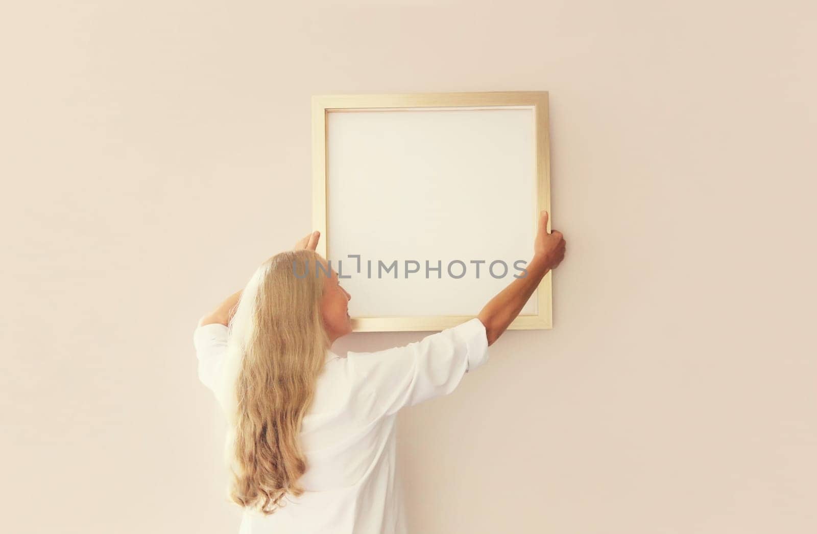 Woman decorating interior, hanging white blank photo frame, wooden picture, artwork mockup on wall in new house