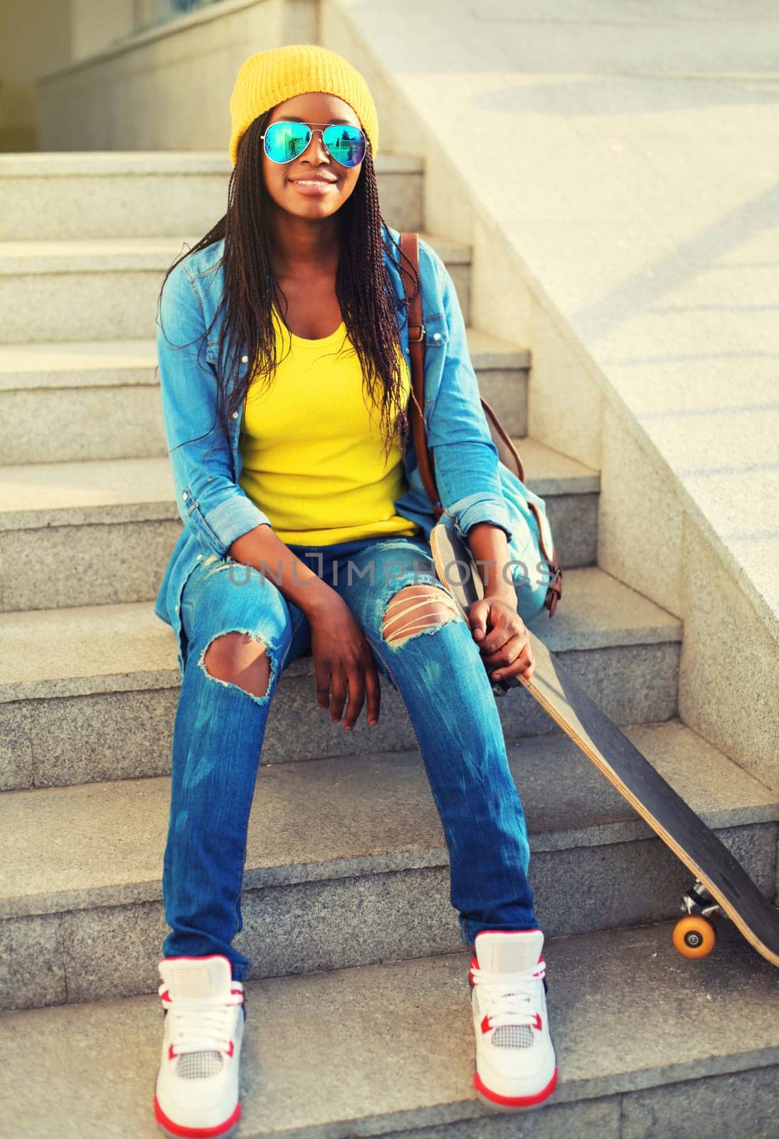 Portrait of stylish happy modern young african woman in colorful bright clothes with skateboard on city street