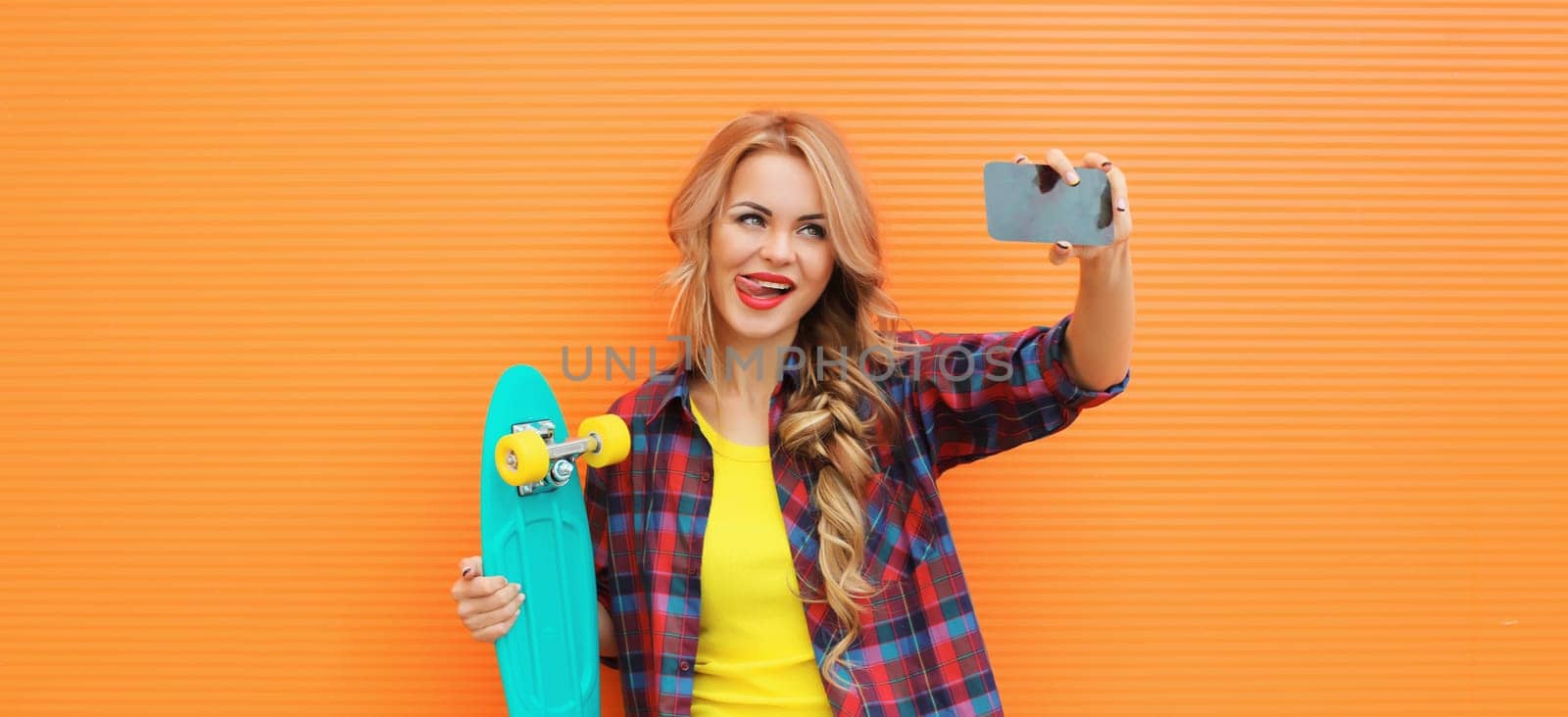 Summer portrait of happy smiling blonde young woman taking selfie with smartphone and skateboard on colorful orange background