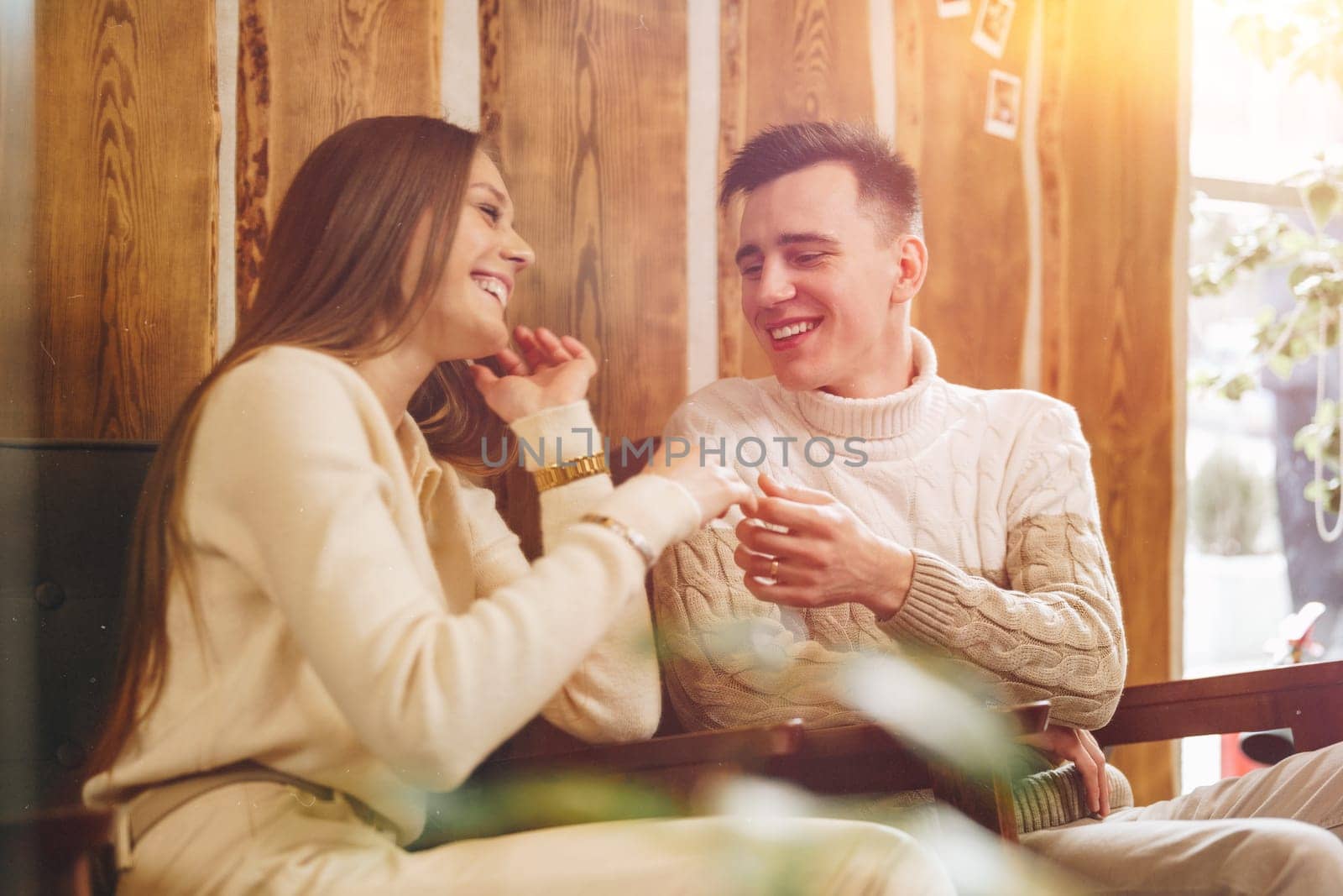 A young man and woman share a joyful moment while sitting at a wooden table in a cafe with a warmly lit interior. They are both smiling and seem to be engaged in a relaxed and friendly conversation. Natural light streams in from the side, highlighting their casual clothing and the intimate setting.