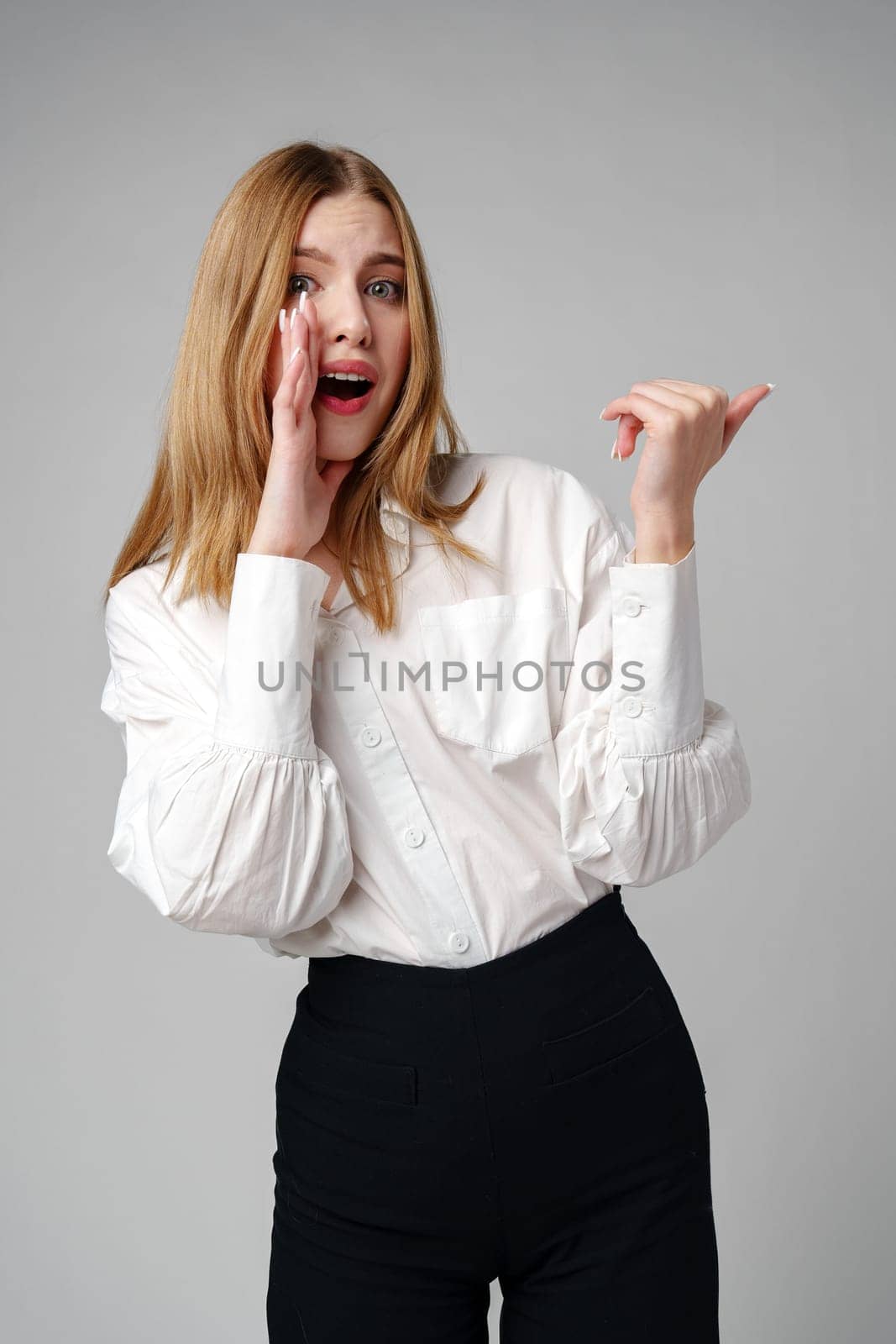Young blonde woman in formal outfit pointing to the side against gray background in studio