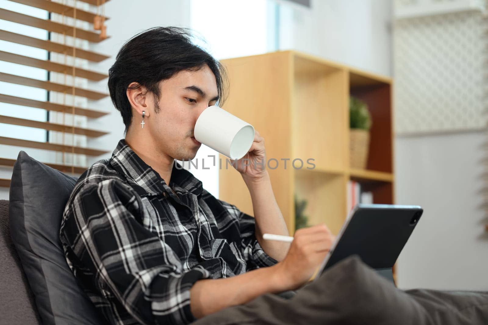 Young creative male freelancer drinking coffee and working on digital tablet at home by prathanchorruangsak