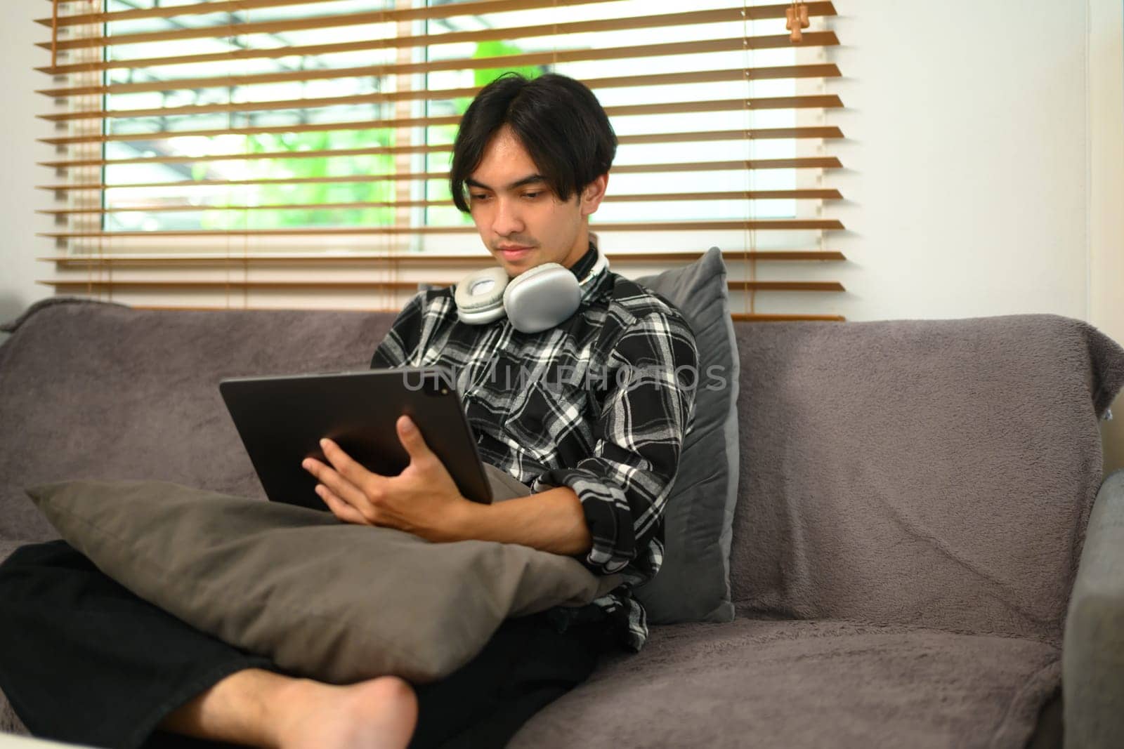 Relaxed young hipster man with headphone using digital tablet on couch at home by prathanchorruangsak