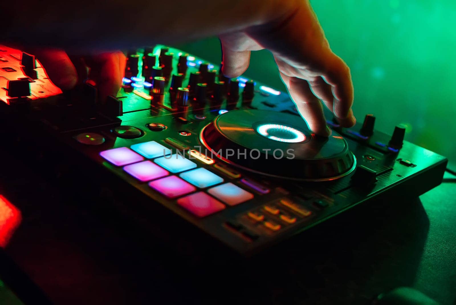 mixer and DJ booth in the nightclub with the arms of the DJ managing the music with colorful lighting and a blurred green background
