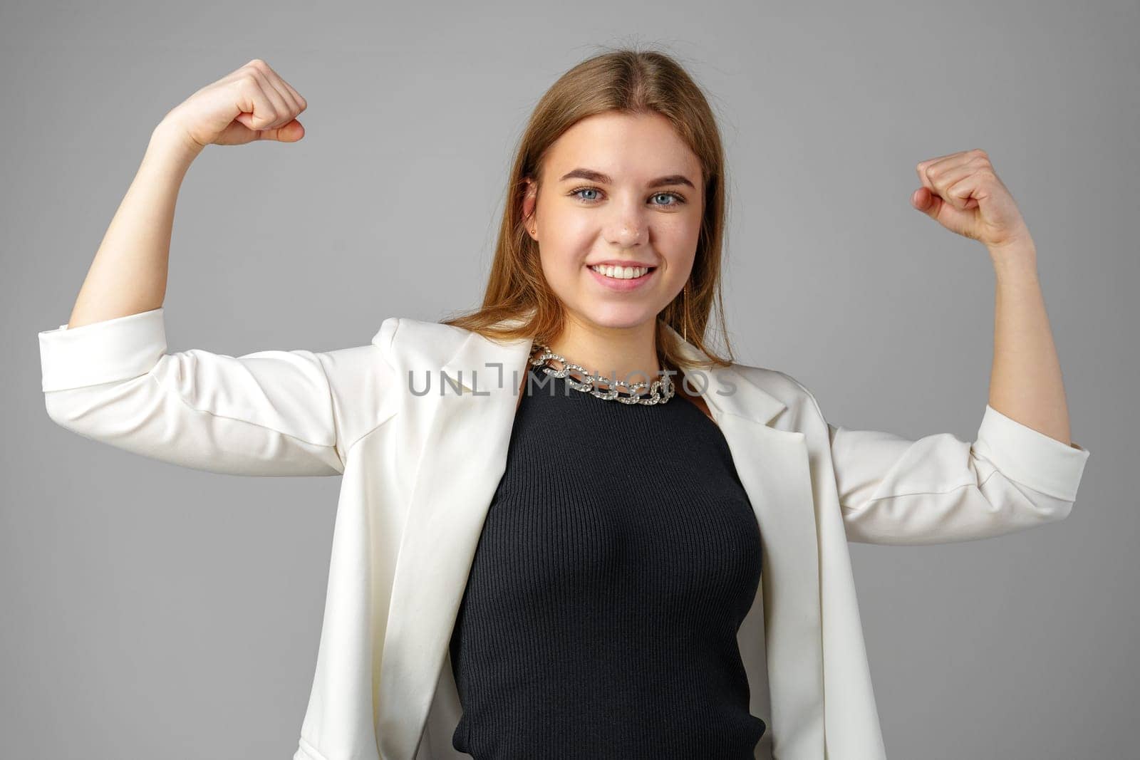Confident Young Businesswoman Flexing Muscles in Smart Casual Attire Against Grey Background by Fabrikasimf