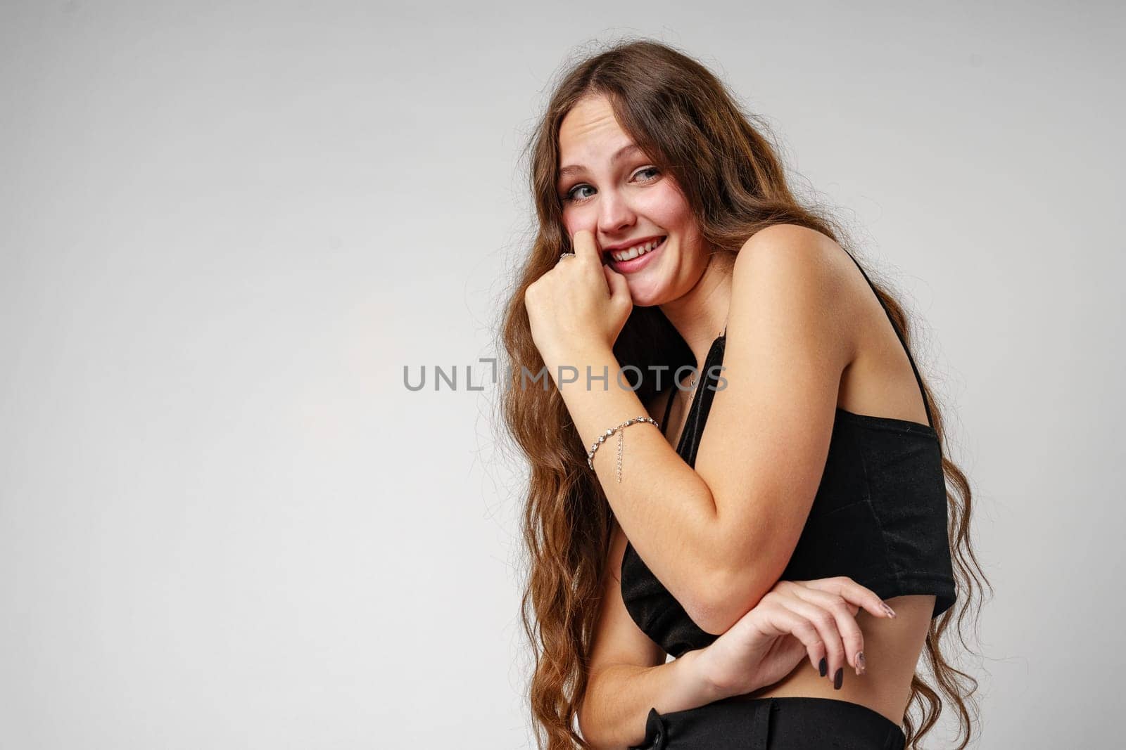 A young woman with long, wavy hair is captured posing expressively with her hand on her cheek and a broad, engaging smile. She is wearing a sleeveless black top which contrasts against a simple, grey backdrop. Her lively expression suggests a moment of genuine happiness or amusement.