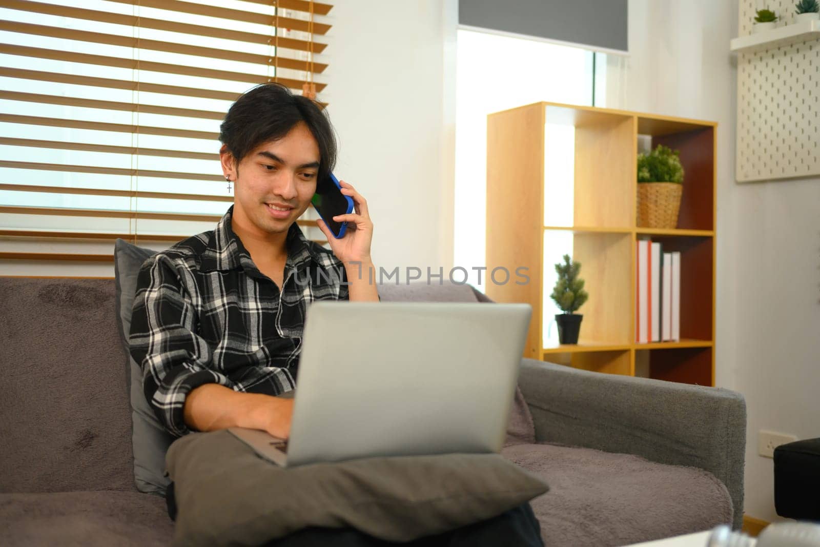 Smiling young man having phone conversation and working with laptop at home by prathanchorruangsak