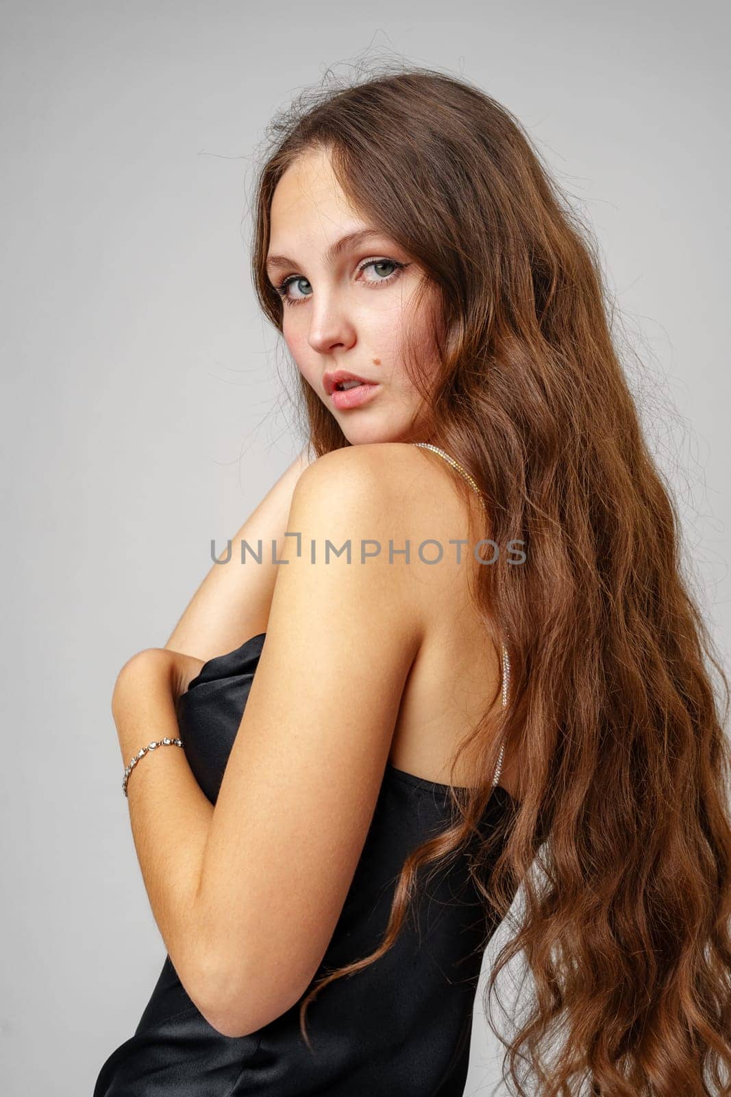 A young woman with long, flowing brown hair gazes over her shoulder with striking blue eyes. She is wearing a strapless black dress, showcasing her bare shoulders, accented by a simple bracelet on her wrist. Her confident yet graceful posture adds to her elegant appearance.