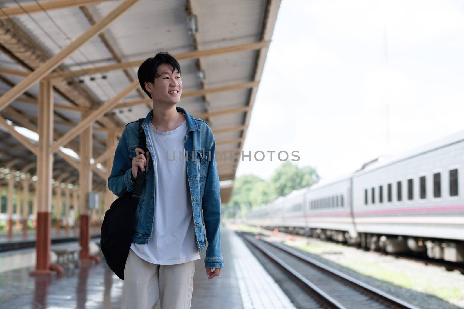 A young man carries a backpack and waits for the train at the train station to travel. by wichayada