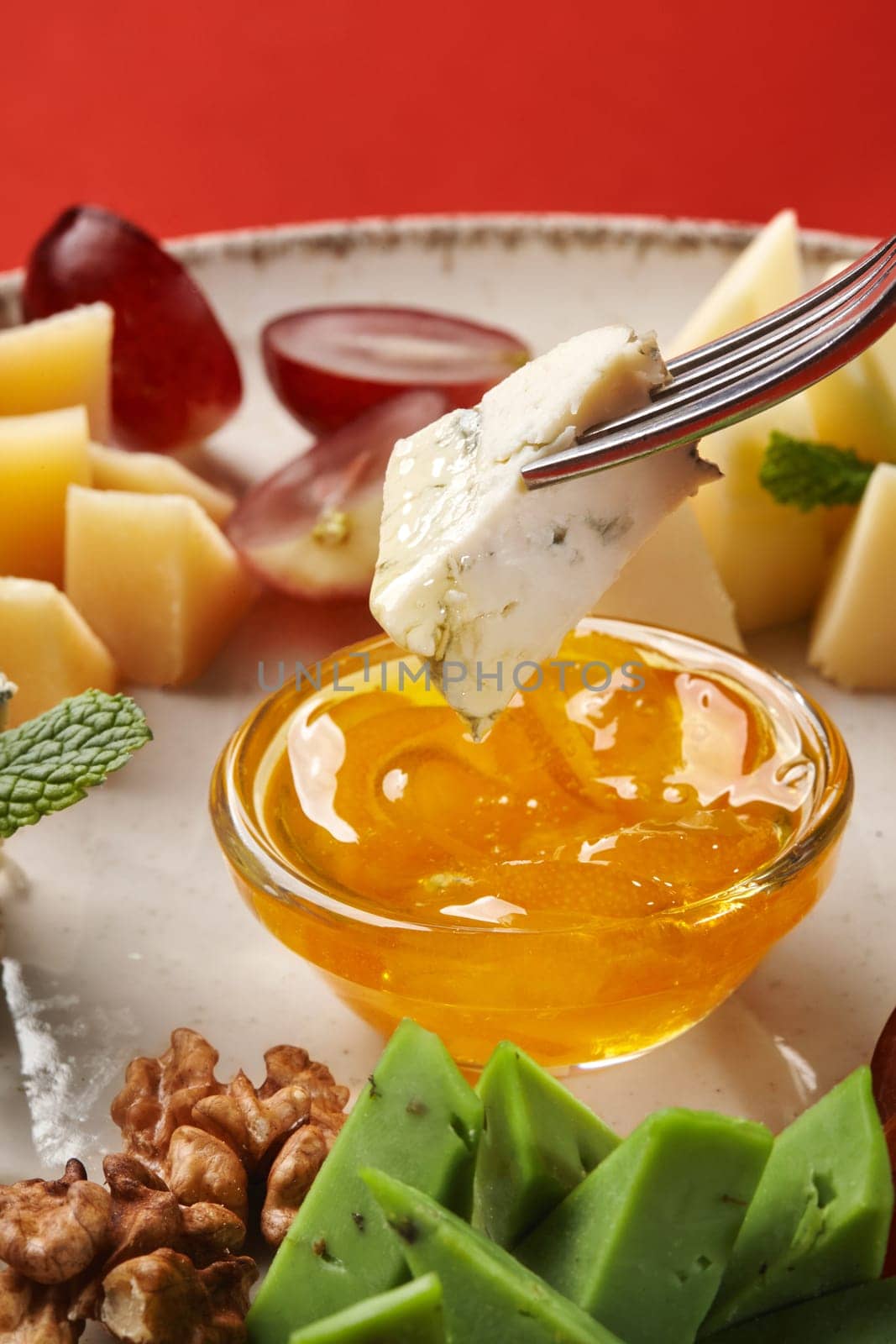 Close-up of piece of blue cheese on fork dipped into bowl of clear honey, served as part of gourmet cheese platter with grapes and walnuts on red background