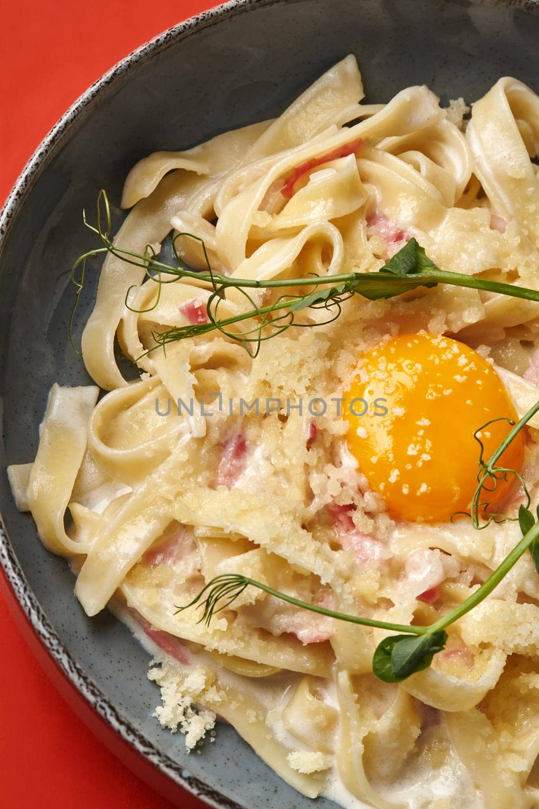 Bowl of traditional Italian pasta carbonara with cured pork pieces, topped with sunny-side up egg, sprinkled with parmesan, and garnished with delicate greens on red background