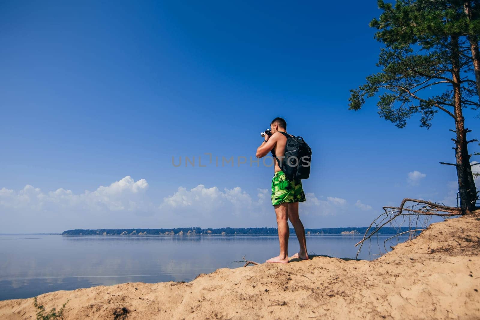 traveler photographer with backpack taking pictures of summer at the river under a blue clean sky on the beach from behind