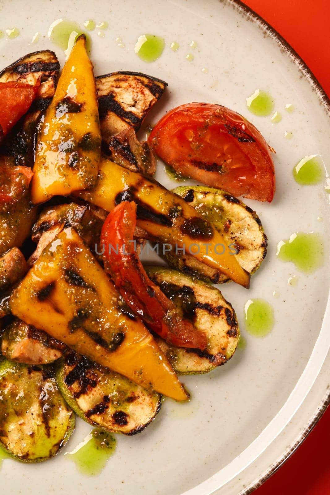 Closeup view of slices of browned grilled zucchini, eggplant, bell pepper, tomato and mushrooms drizzled with spicy herb oil, served on ceramic plate against red background