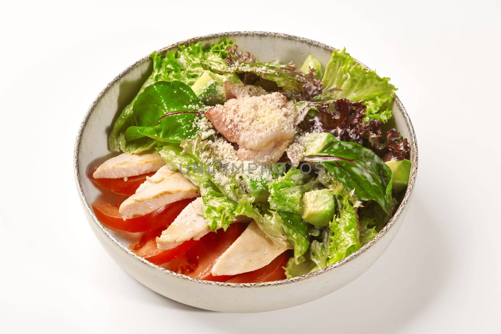 Rustic ceramic bowl of fresh salad with grilled chicken strips, tomato slices, ripe avocado and mixed greens seasoned with creamy Caesar dressing and grated Parmesan cheese. Perfectly balanced dinner