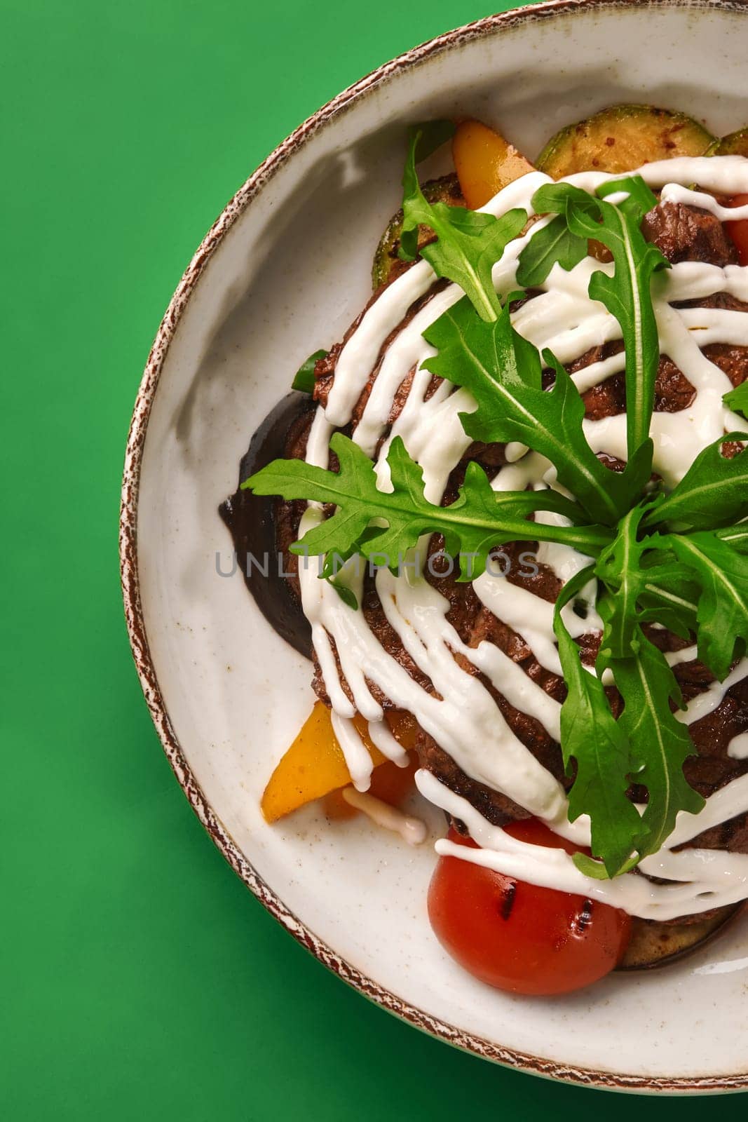 Comforting warm salad with grilled tender slices of beef and vegetables drizzled with creamy dressing, topped with fresh aromatic arugula, served in ceramic bowl on green backdrop